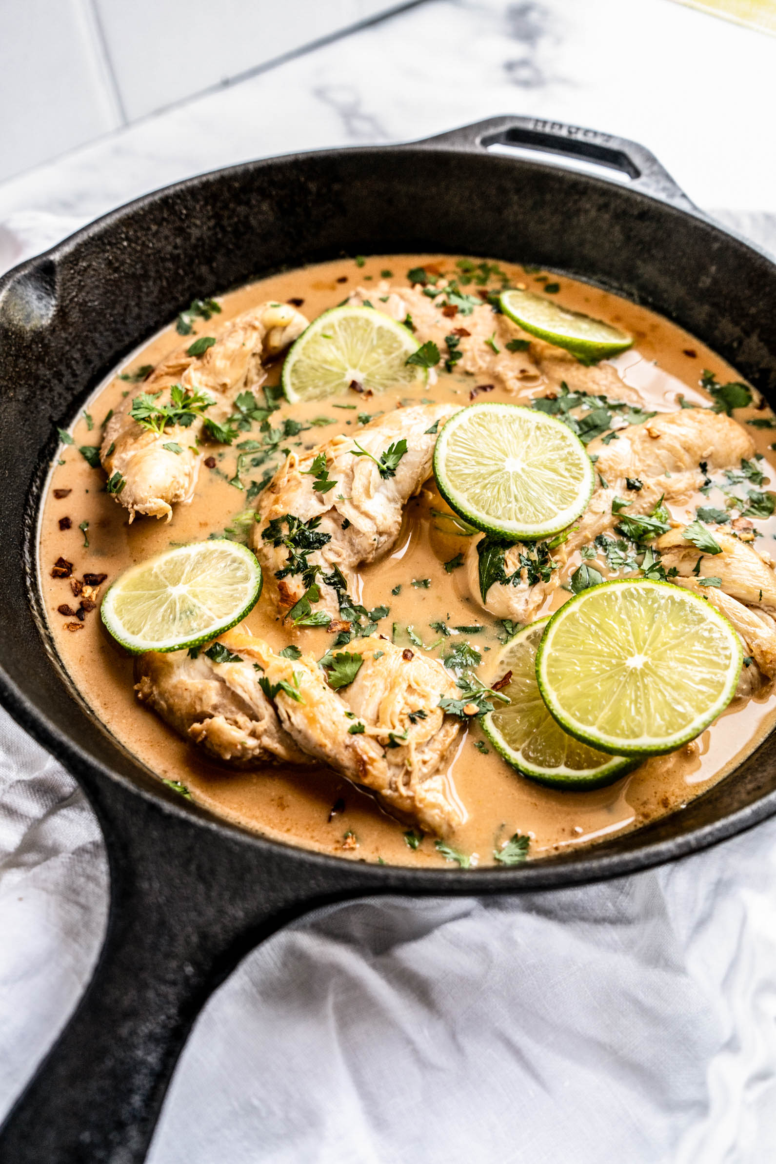 One pan coconut lime chicken simmered in a creamy coconut sauce with tender chicken, fresh lime slices, cilantro, and red pepper flakes in a cast iron skillet on a light marble background.
