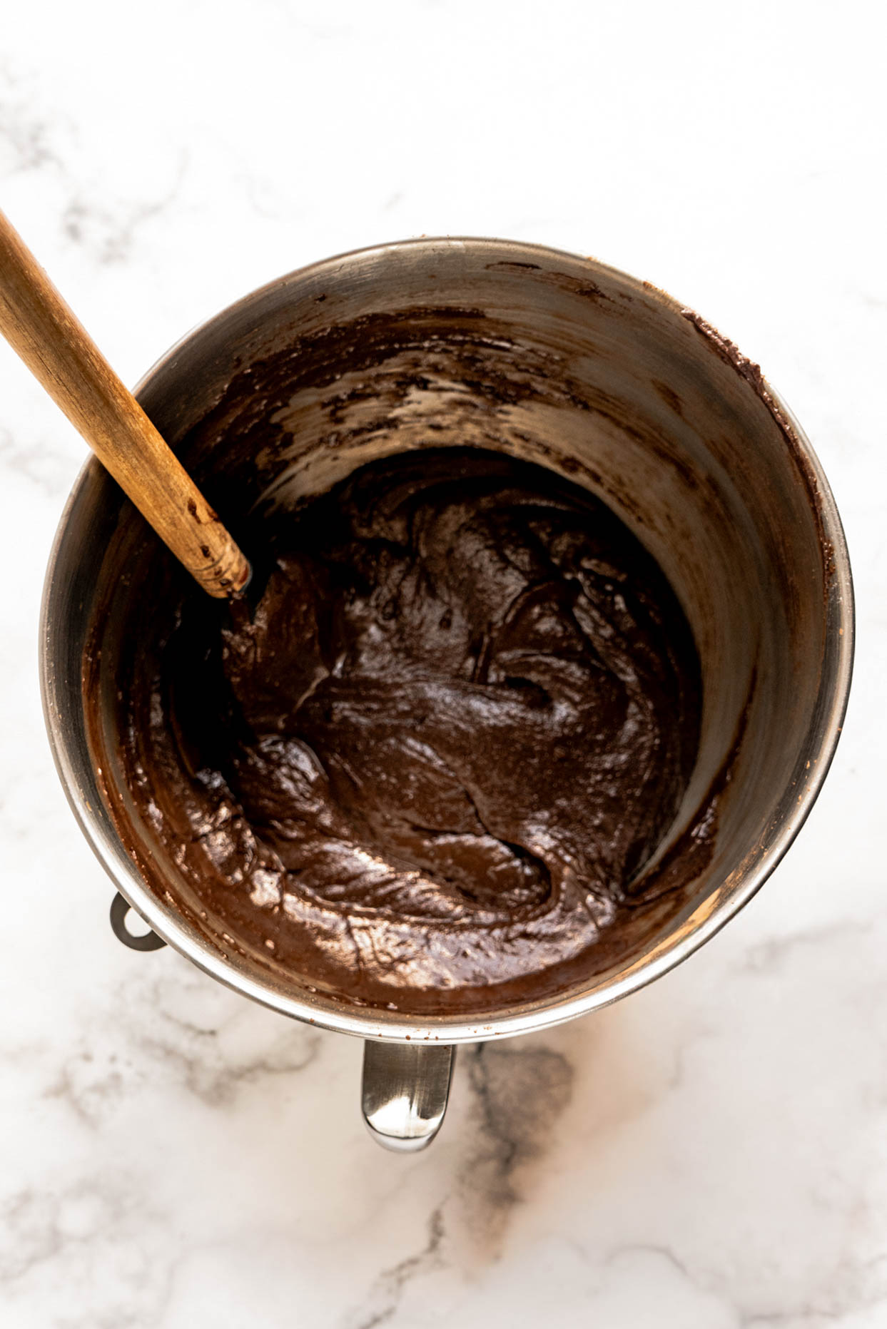 Rich chocolate brownie batter made with peanut butter whiskey being mixed in a metal bowl with a wooden spoon on a marble countertop.