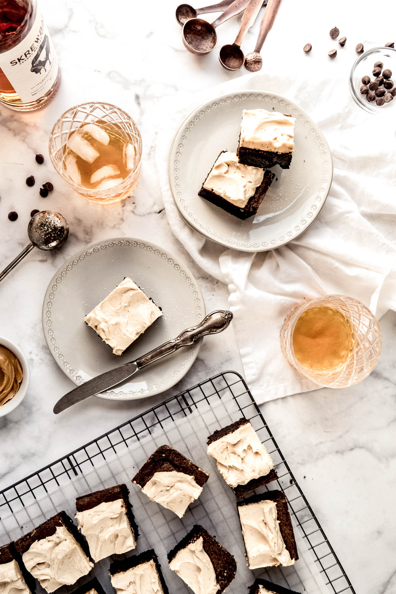 Overhead entertaining scene with fudgy brownies topped with peanut butter whiskey frosting on plates and a cooling rack, served alongside glasses of peanut butter whiskey on a marble table.