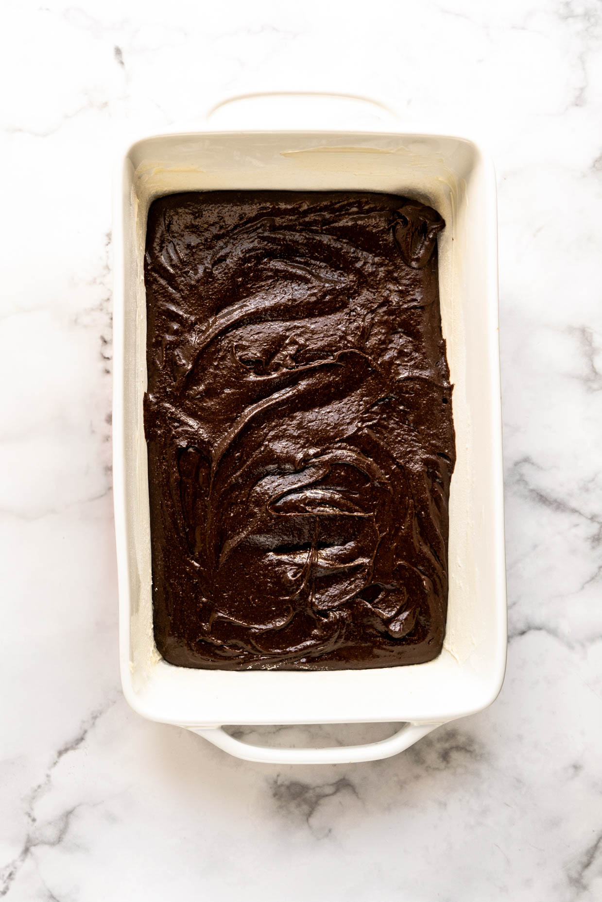 Chocolate brownie batter made with peanut butter whiskey spread evenly in a white baking dish on a marble surface, ready to bake.