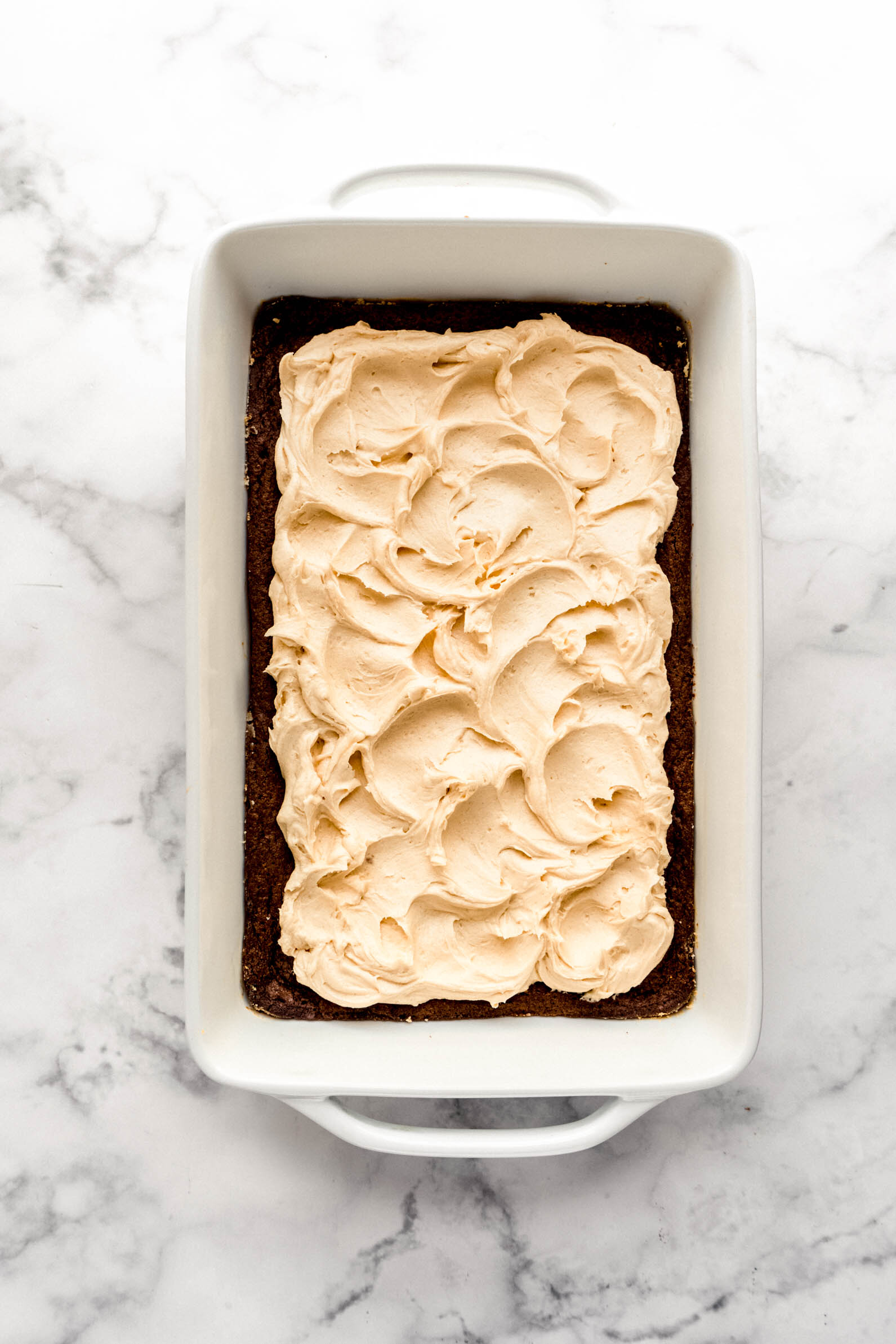 Fudgy chocolate brownies in a white baking dish topped with thick swirls of creamy peanut butter whiskey frosting on a white marble surface.