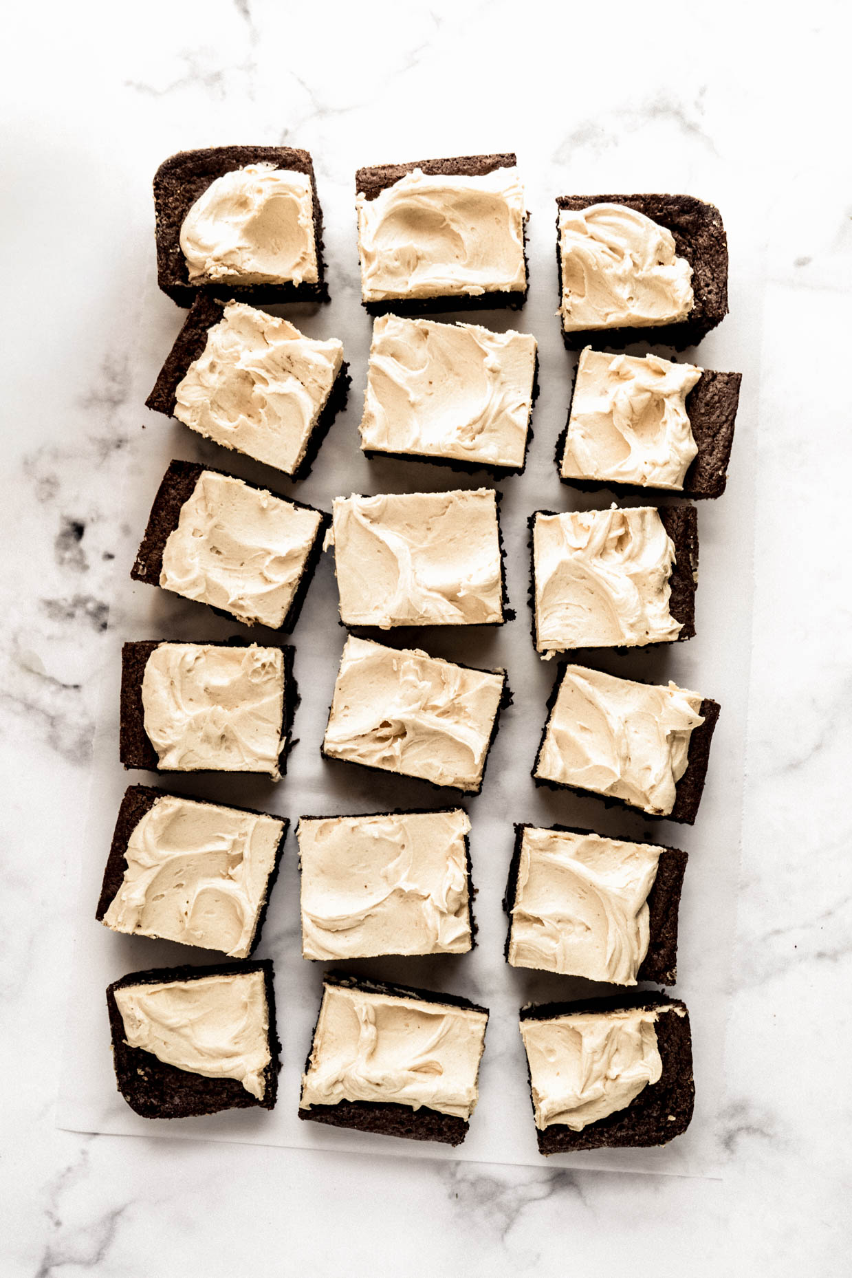 Overhead view of fudgy chocolate brownies topped with smooth peanut butter whiskey frosting arranged in neat rows on parchment paper over a white marble background.
