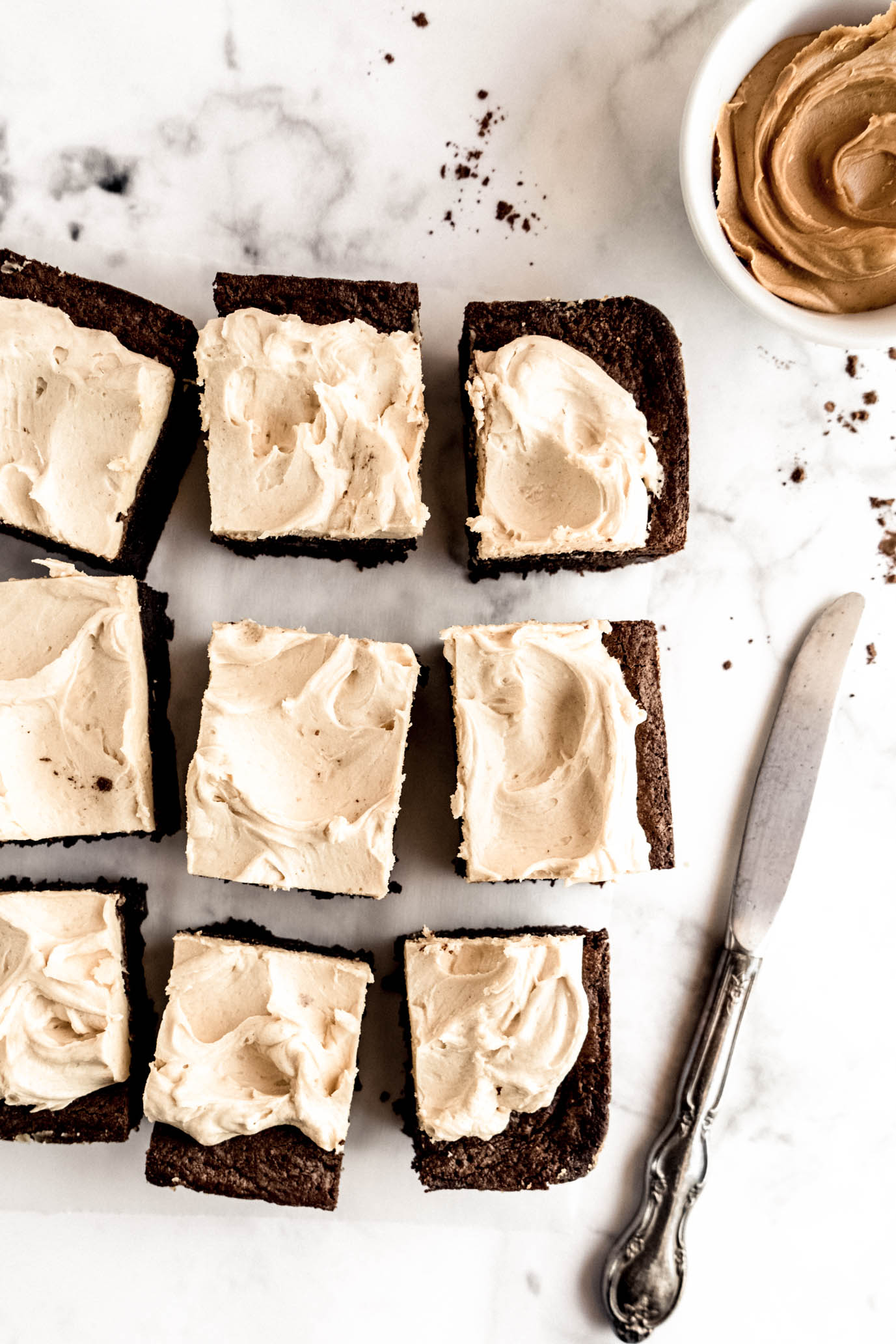 Overhead view of fudgy chocolate brownies topped with creamy peanut butter whiskey frosting arranged on a white marble surface, with a small bowl of peanut butter whiskey frosting and a butter knife beside the brownies.