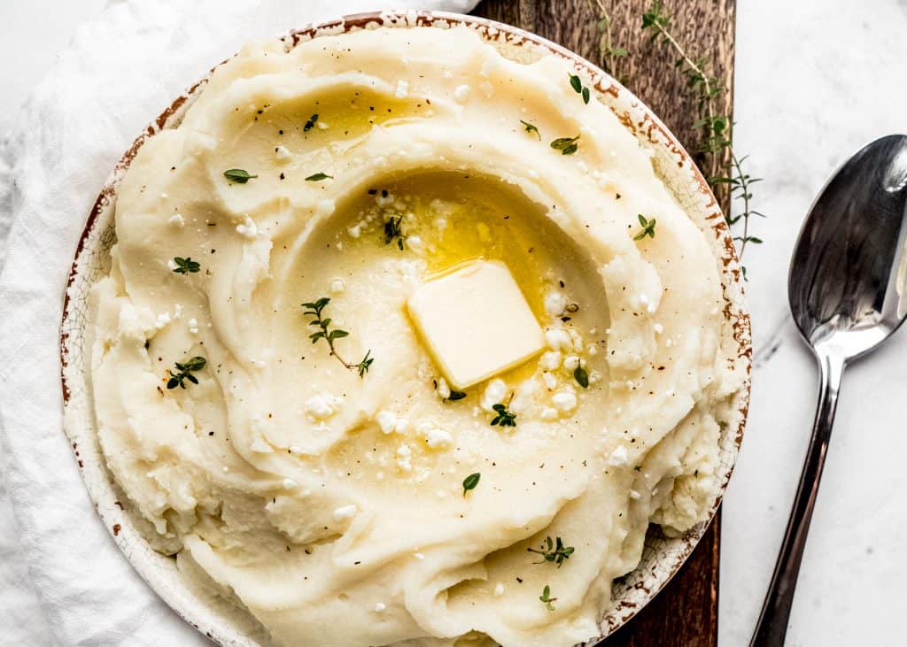 goat cheese mashed potatoes in serving bowl with butter and fresh thyme on top. The bowl is sitting on top of a wood cutting board with a serving spoon to the right