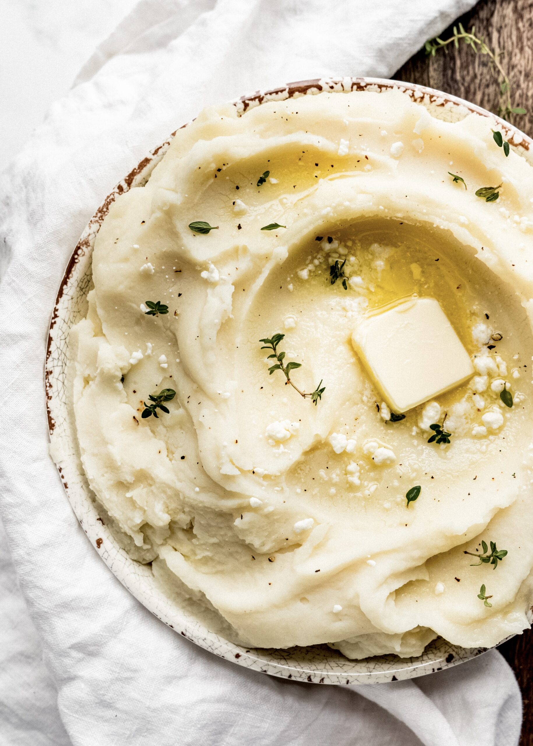 goat cheese mashed potatoes in serving bowl with butter and fresh thyme on top. The bowl is sitting on top of a wood cutting board