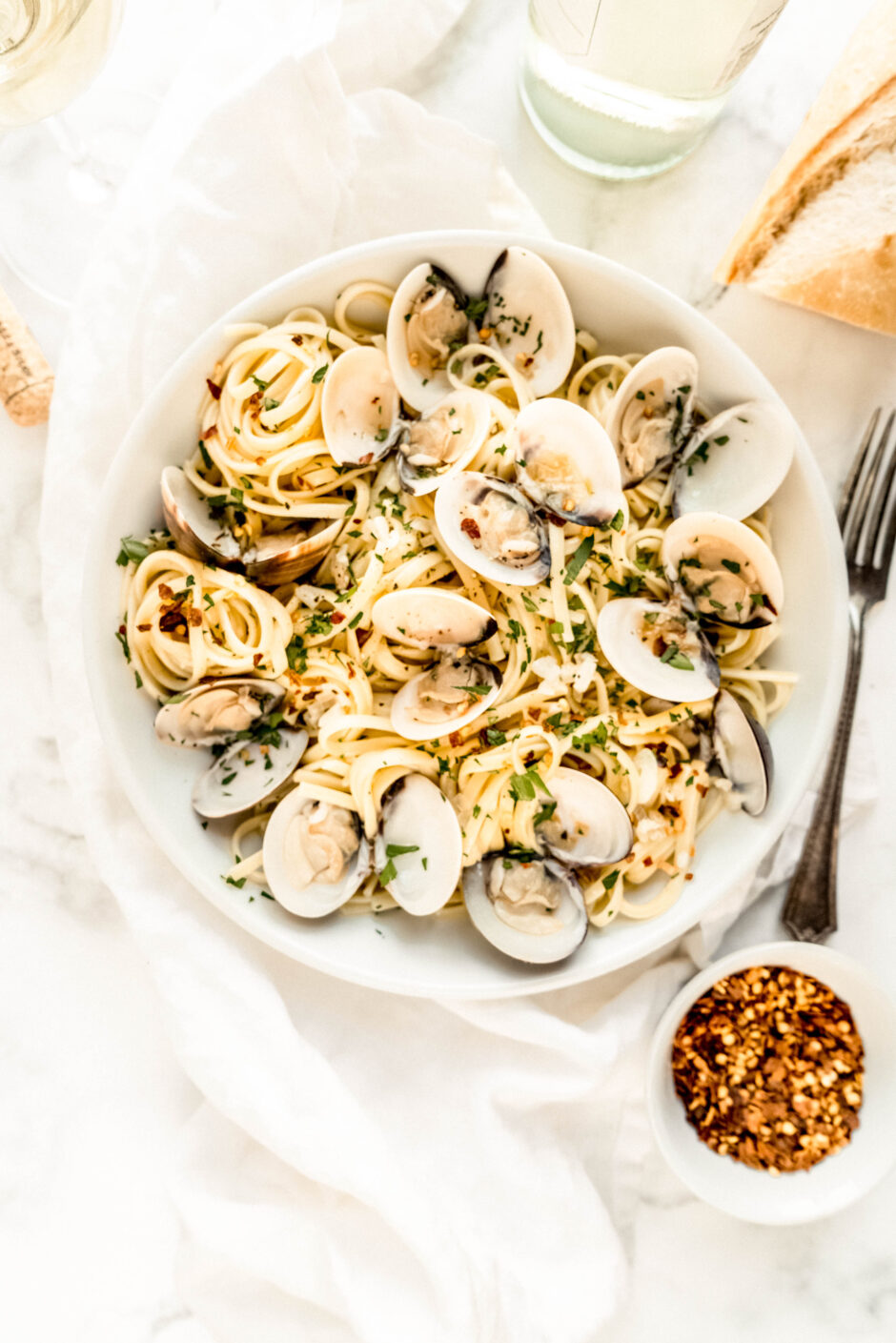 Overhead view of linguine with clams in white wine sauce, featuring tender pasta and opened clams tossed with garlic, olive oil, and lemon, finished with fresh parsley and red pepper flakes on a white linen backdrop.