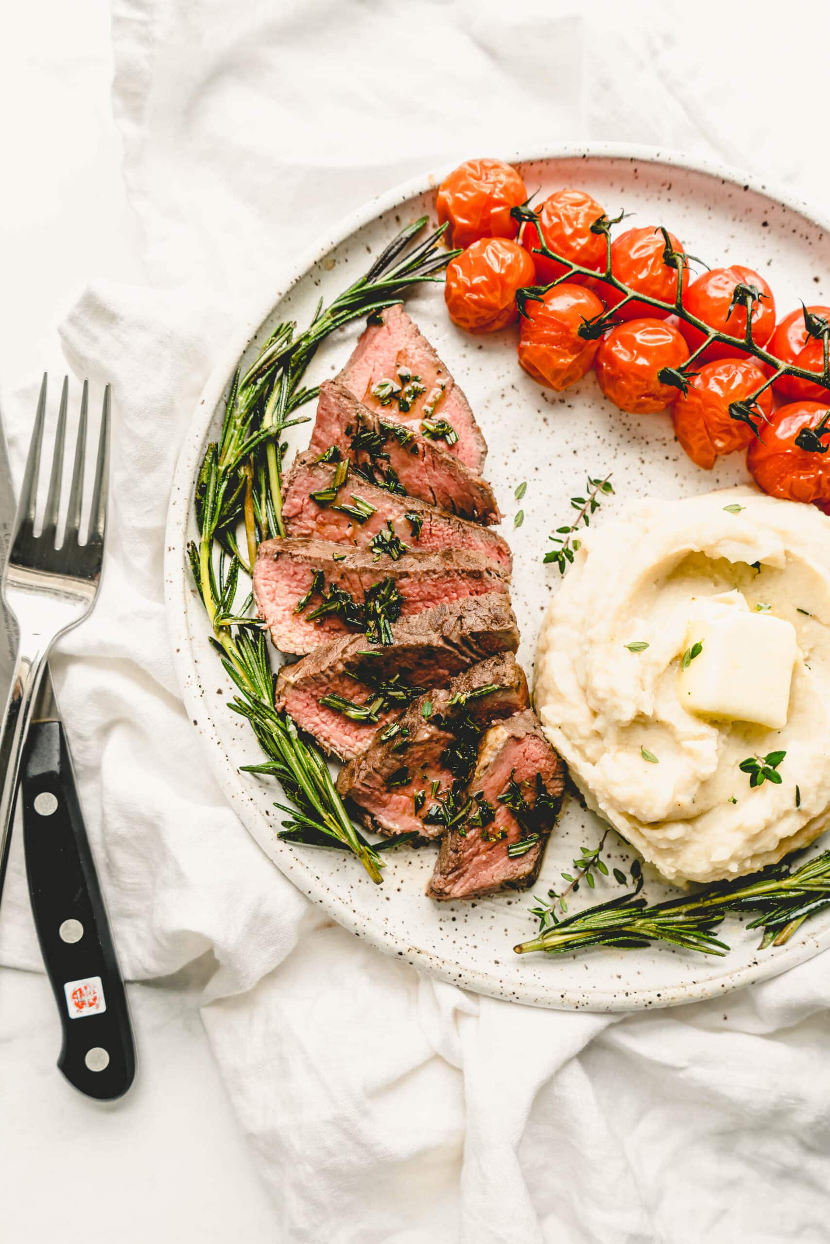 Sous vide steak sliced medium-rare and topped with fresh herbs, served on a speckled ceramic plate with creamy mashed potatoes, roasted cherry tomatoes on the vine, and sprigs of rosemary on a light marble backdrop.