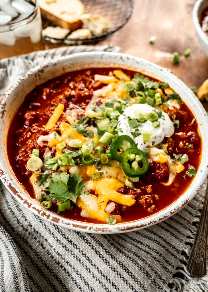 A 45 degree angle shot of a bowl of turkey chili garnished with shredded cheese, cilantro, sliced green onion and sour cream. The bowl is sitting on a striped napkin on a wood table.