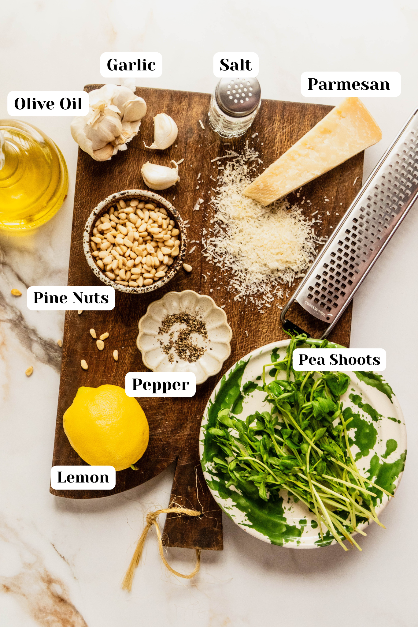 Bright overhead ingredient shot for pea pesto on a wooden board over a white marble background, featuring fresh pea shoots in a bowl, a wedge of parmesan cheese with grated shavings, garlic cloves, pine nuts, lemon, olive oil, salt, and cracked pepper neatly arranged and labeled.