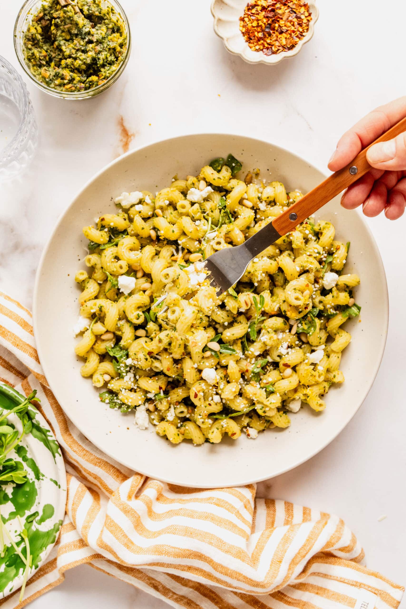 Bright overhead shot of cavatappi pasta tossed in pea pesto, topped with crumbled cheese, pine nuts, and red pepper flakes, with a hand holding a fork lifting a bite on a white plate over a marble background.