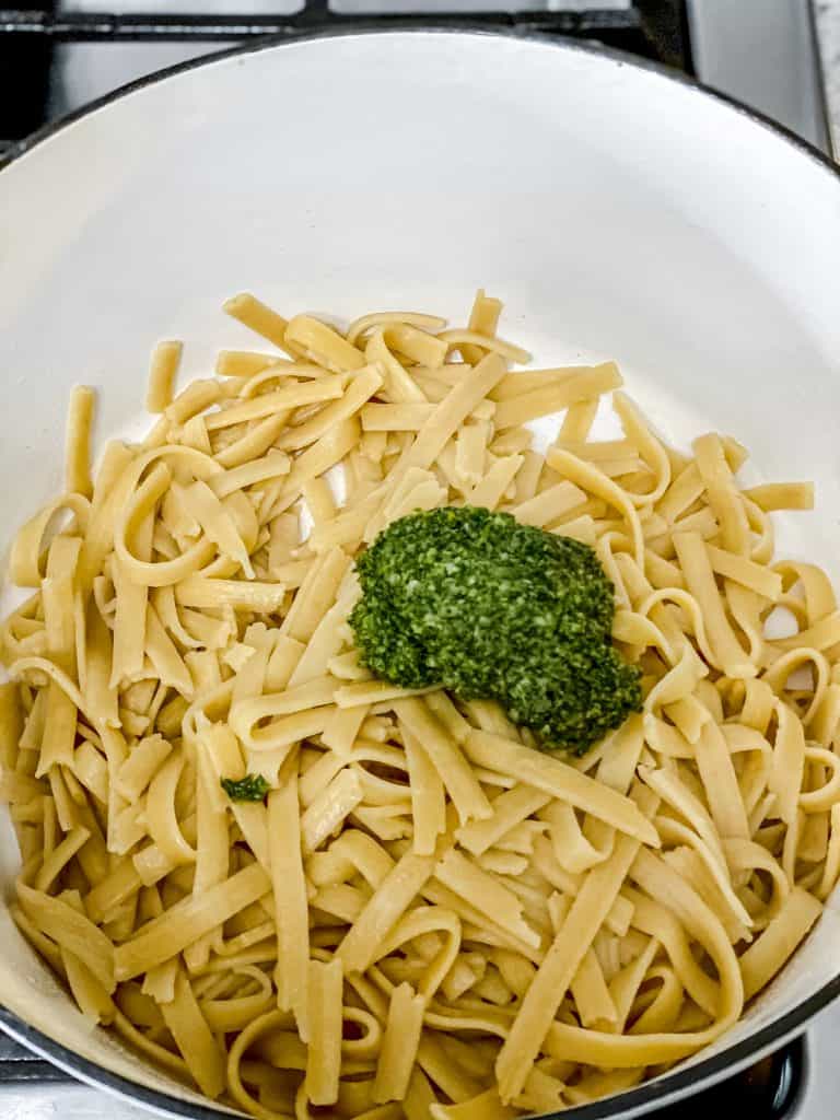 Cooked fettuccine in a white pan topped with a spoonful of pesto, preparing pesto pasta with shrimp before tossing the sauce into the noodles.