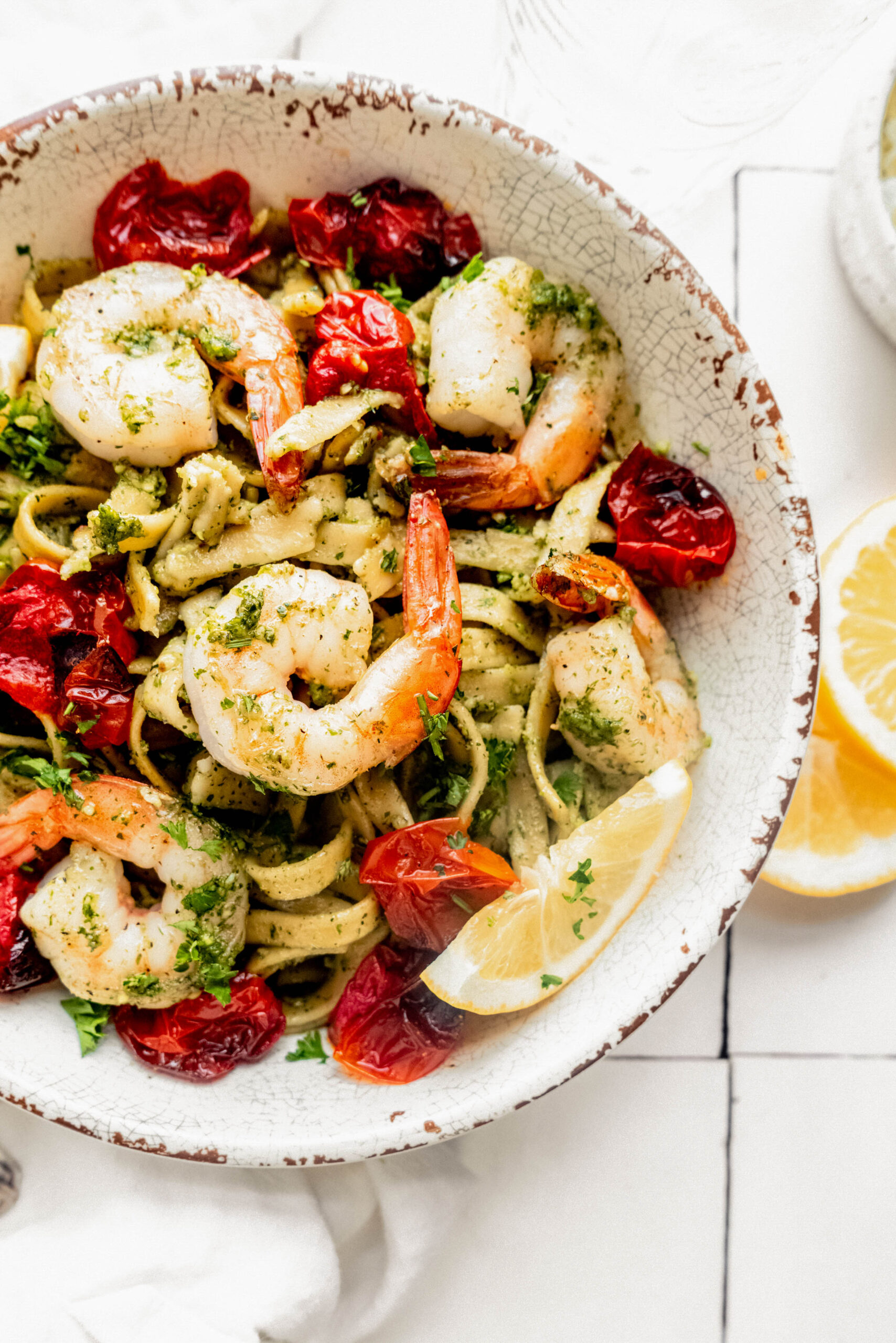 Close up view of pesto pasta with shrimp, roasted cherry tomatoes, and fettuccine in a rustic bowl, garnished with lemon wedges and fresh herbs on a white marble surface.