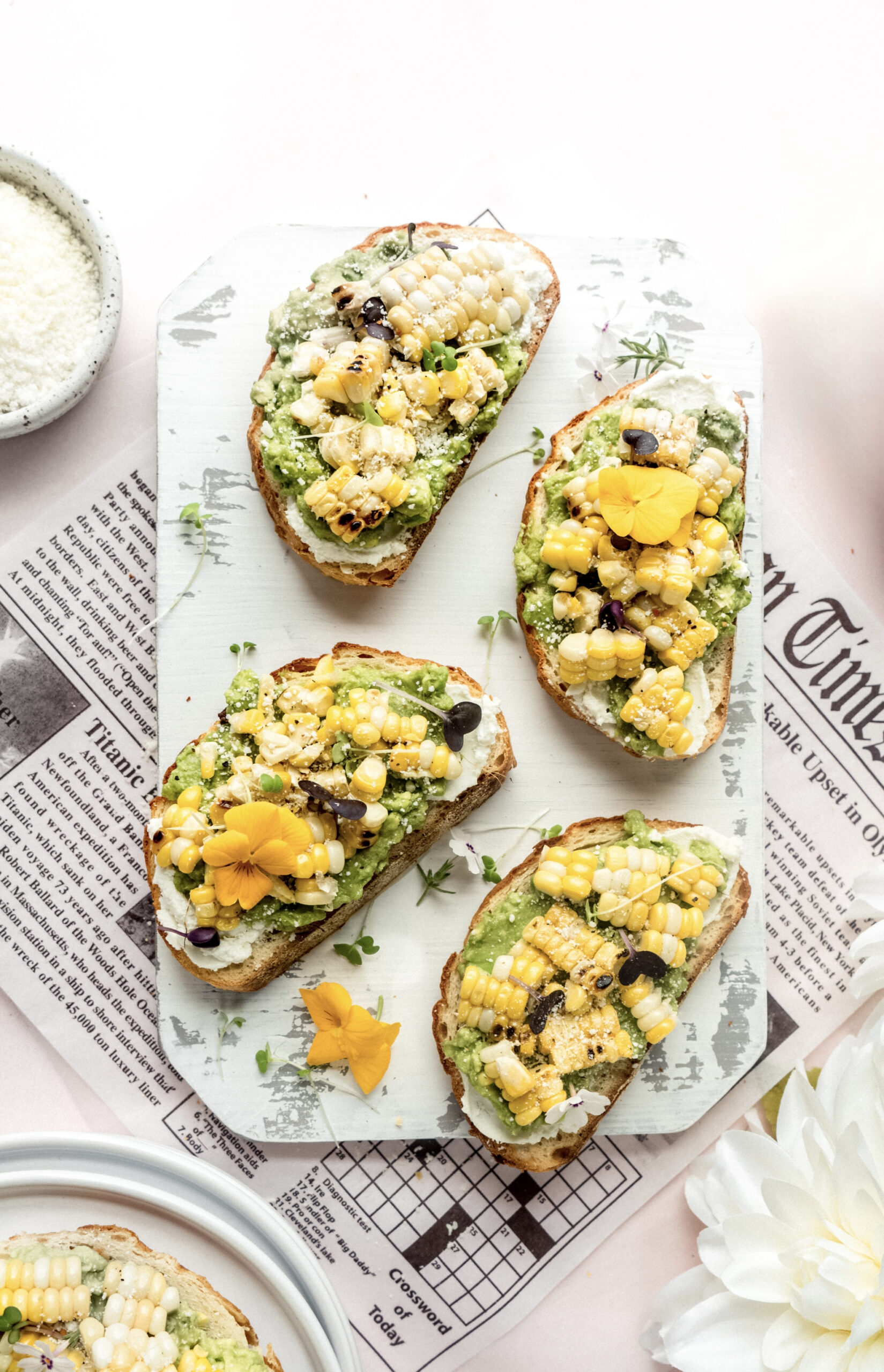 Alt text:
A bright and colorful display of Mexican bruschetta topped with mashed avocado, grilled sweet corn, crumbled cheese, and edible flowers on toasted baguette slices, arranged on a white serving board over newspaper.