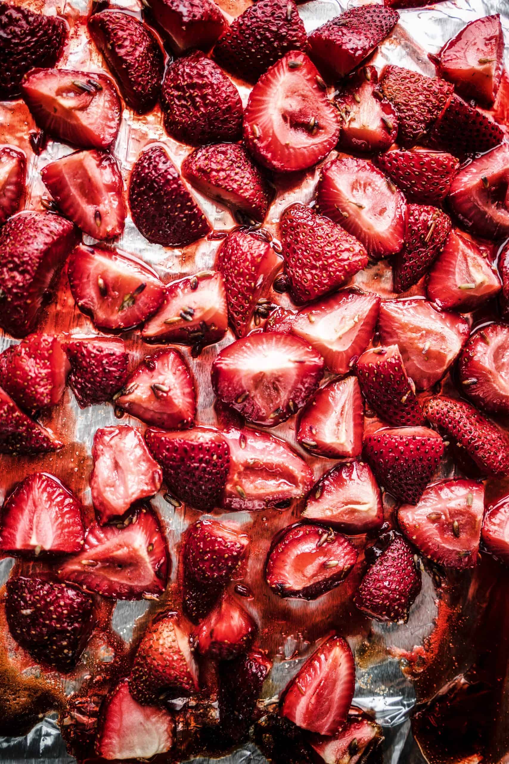 Close-up image of roasted strawberries glistening with syrup on a foil-lined baking sheet, prepared for strawberry bruschetta. The berries are caramelized and slightly softened, showing their vibrant red color and juicy texture, perfect for layering onto toasted bread with a creamy spread.