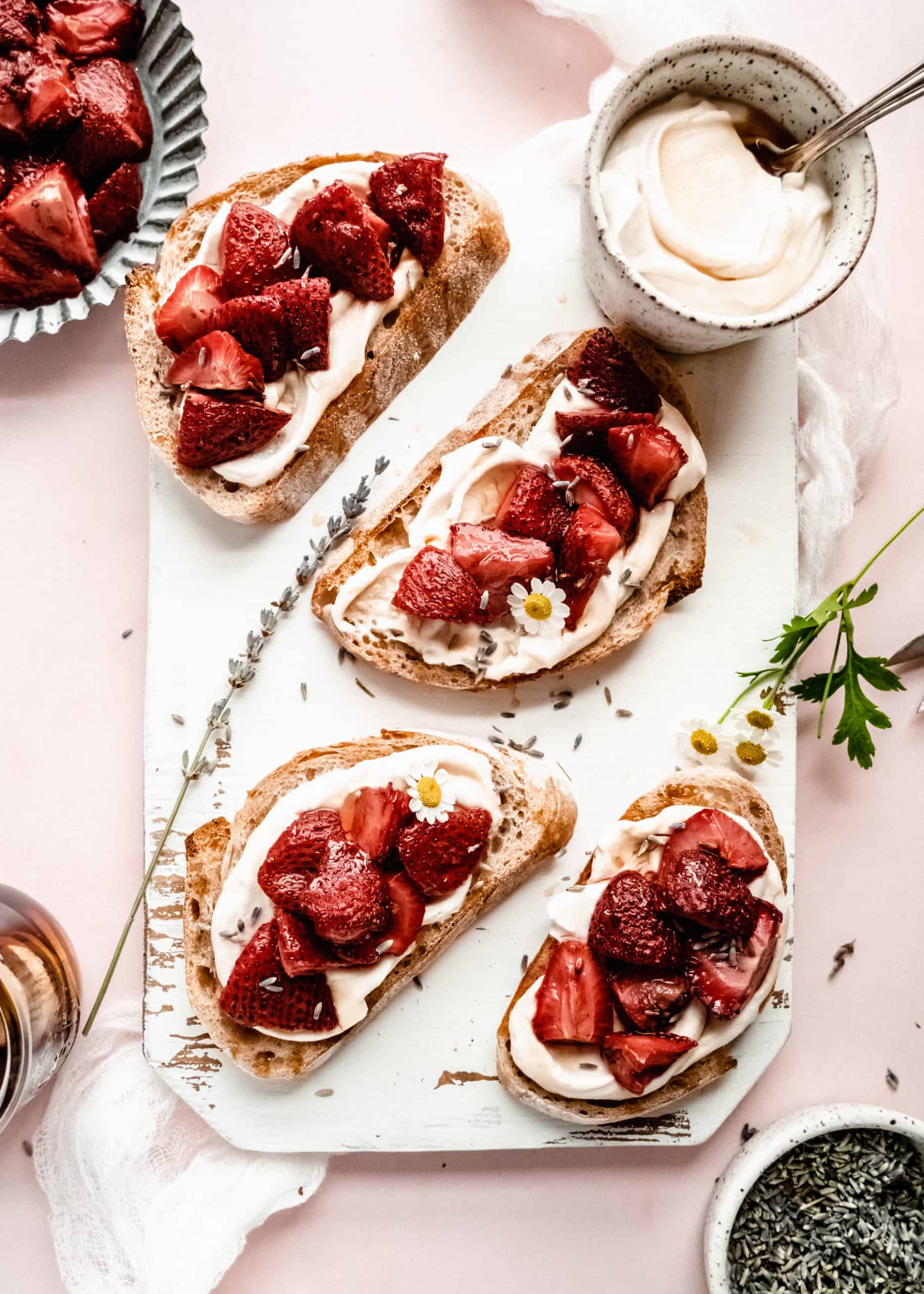 Overhead view of strawberry bruschetta on a white serving board. Each slice of crusty bread is topped with a creamy spread and juicy roasted strawberries, garnished with lavender buds and small edible flowers. A bowl of the creamy spread and a dish of strawberries sit nearby on a soft pink background, creating a bright and elegant presentation.