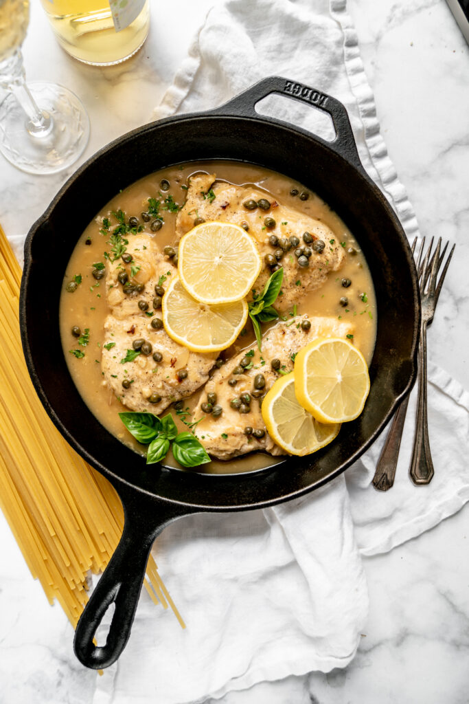 overhead shot of healthy chicken piccata in a cast iron skillet garnished with lemon slices and basil on a white marble background with raw linguine on the side