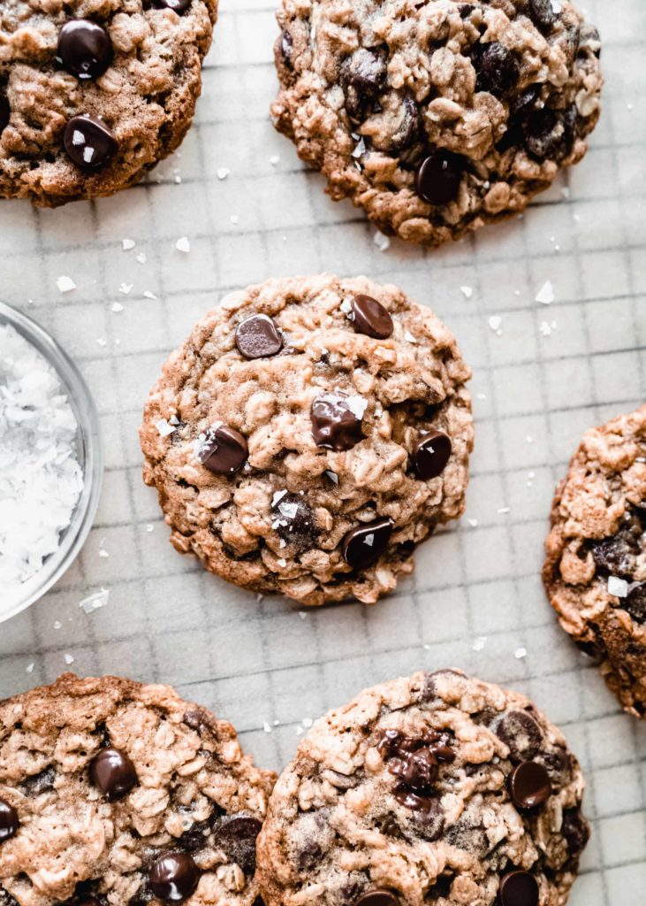 baked Chewy Oatmeal Chocolate Chip Cookies on a parchment lined cooling rack next to a bowl of flaky sea salt.