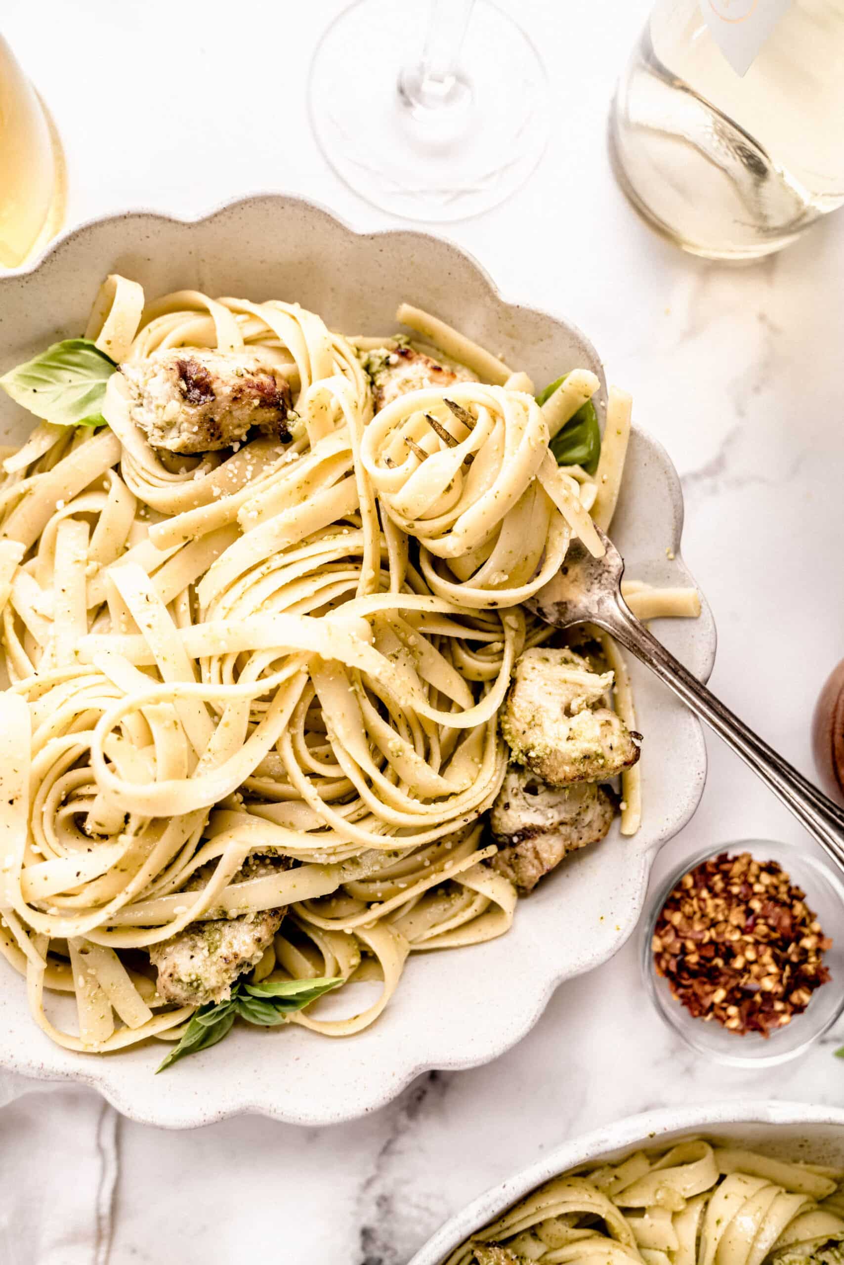 Close-up overhead of chicken pasta pesto with fettuccine-style noodles tossed in basil pesto, juicy chicken pieces, and a light sprinkle of parmesan, served in a scalloped bowl on a marble surface.