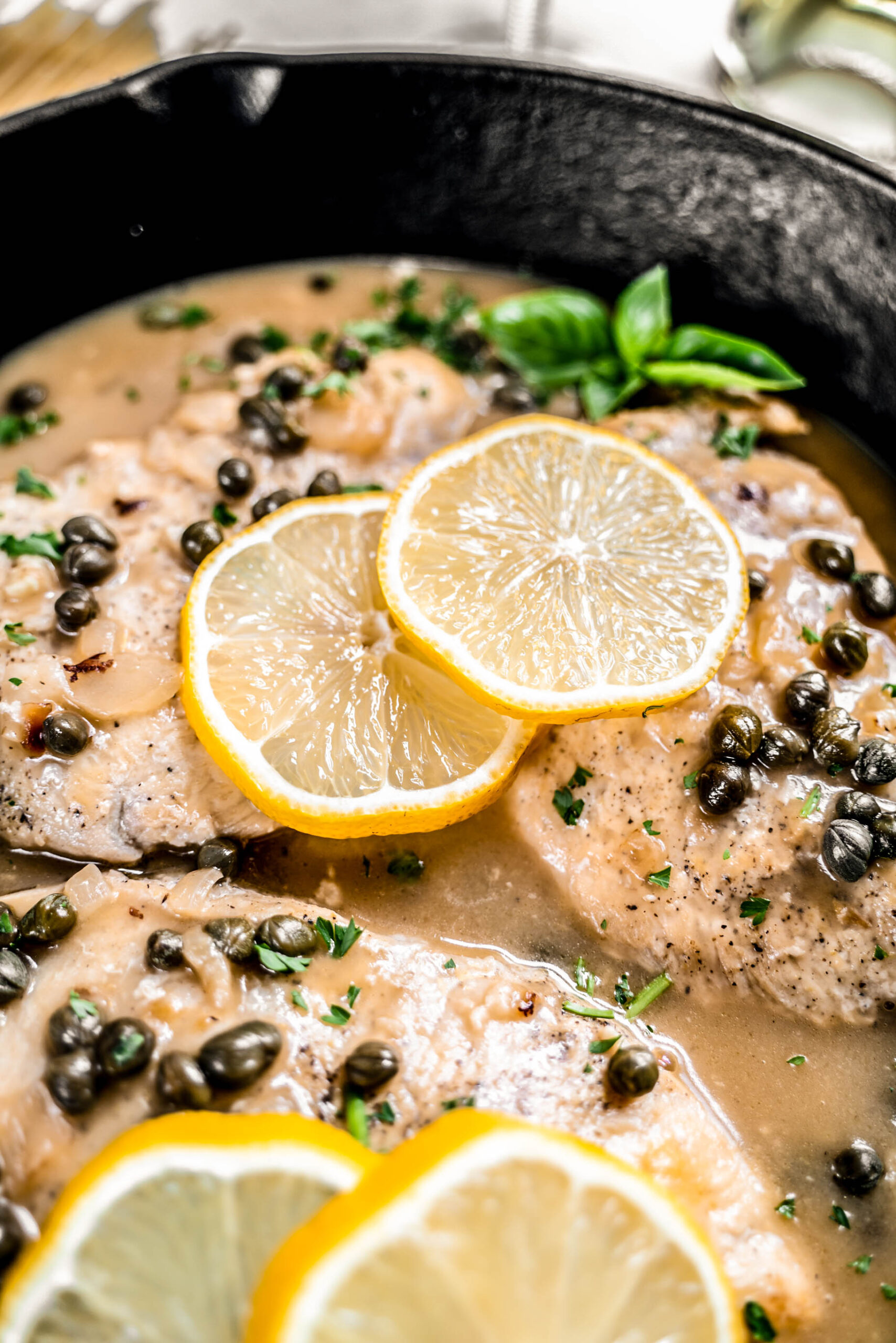 Up close shot of Healthy chicken piccata recipe made with pan-seared chicken breasts simmered in a lemon garlic white wine sauce with capers and shallots, garnished with fresh basil and lemon slices in a cast iron skillet.