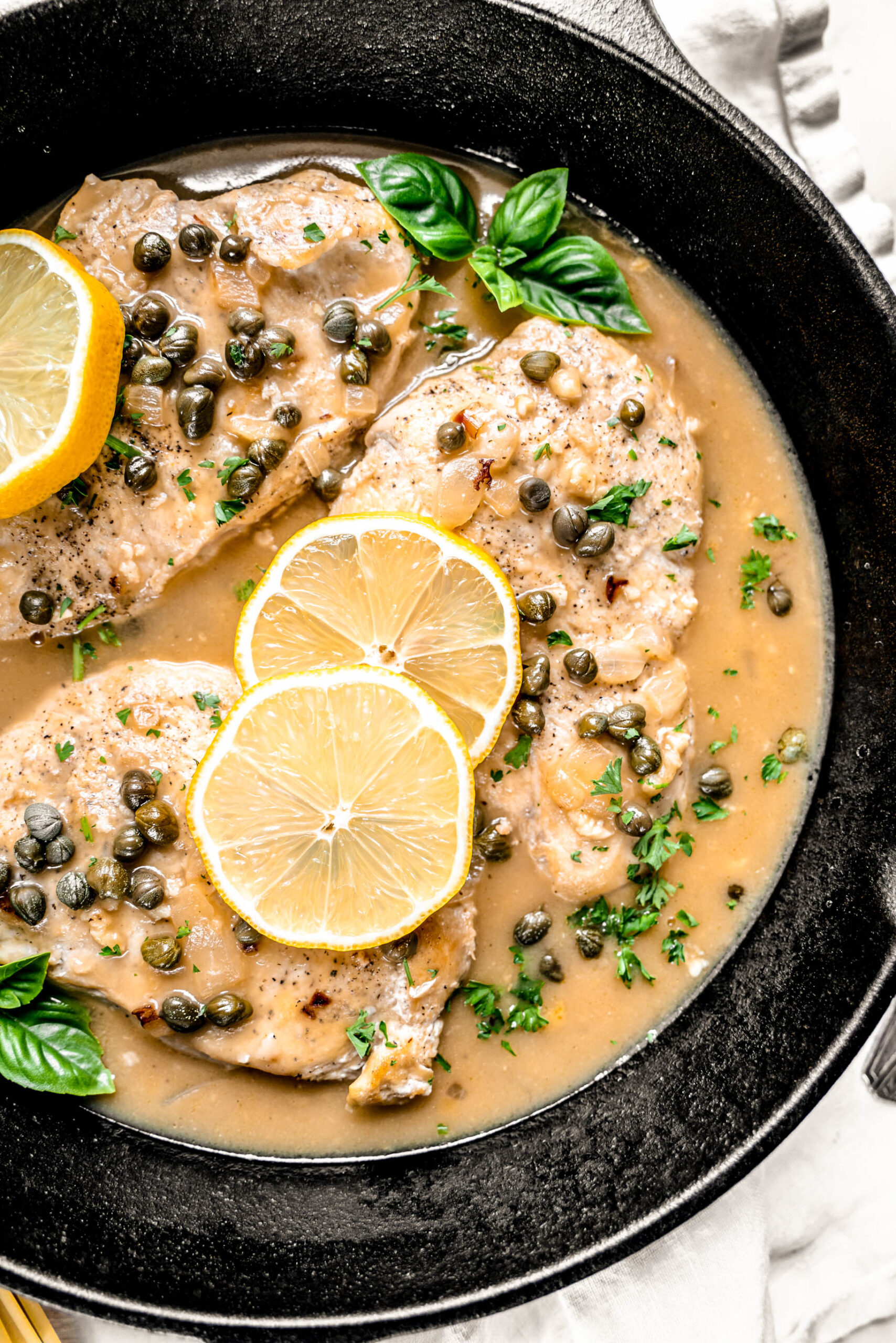 Overhead shot of Healthy chicken piccata recipe made with pan-seared chicken breasts simmered in a lemon garlic white wine sauce with capers and shallots, garnished with fresh basil and lemon slices in a cast iron skillet.
