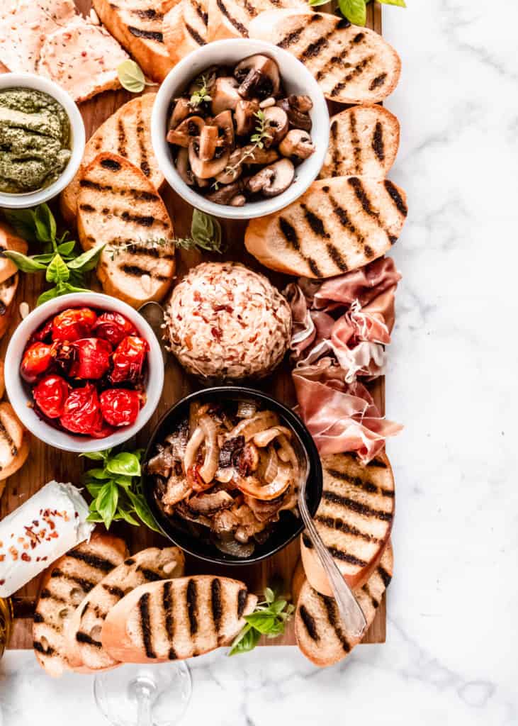 close up of caramelized onions, prosciutto, sauteed mushrooms, and roasted grape tomatoes on board in bowls with grilled bread surrounding