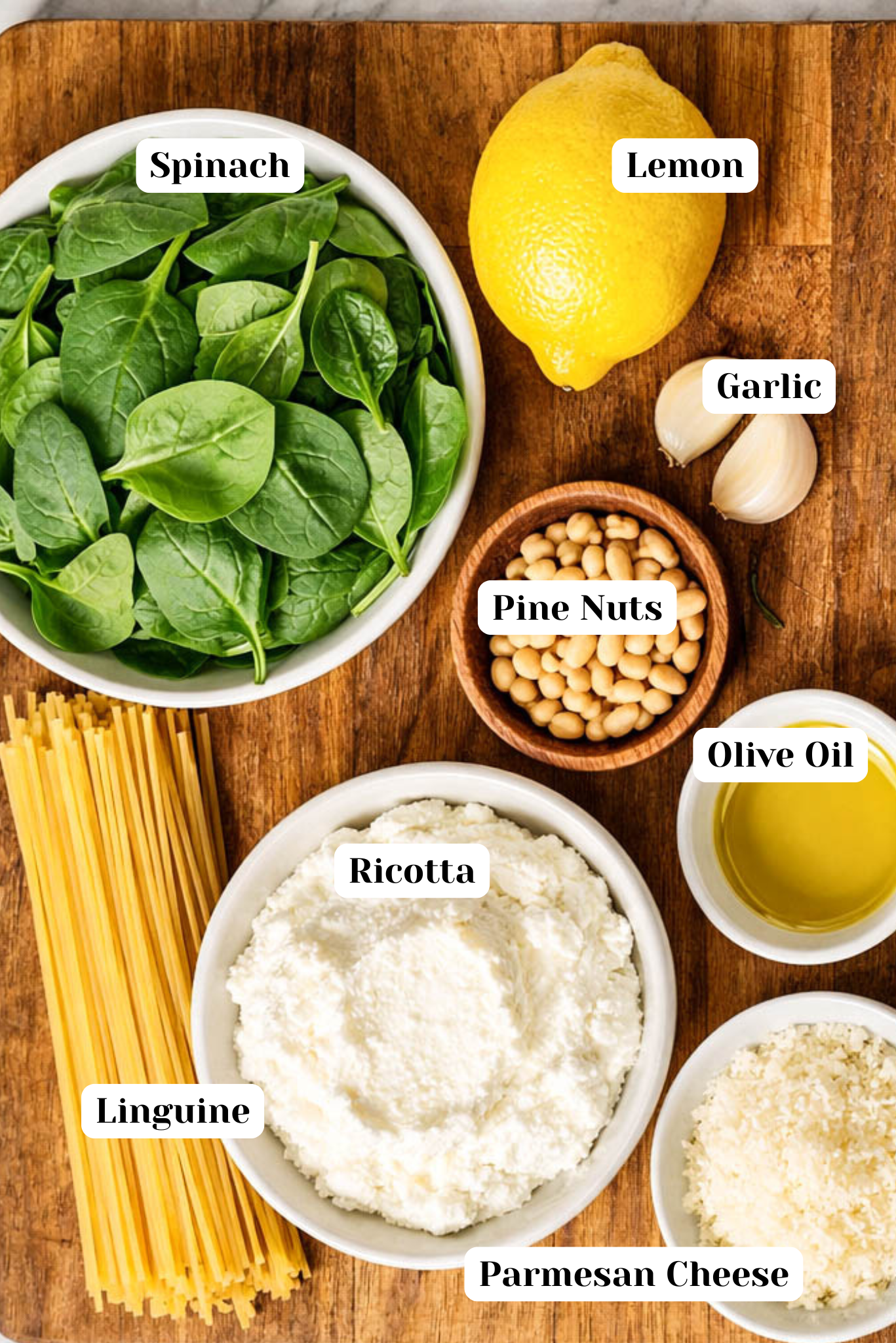 Lemon ricotta pasta recipe ingredients arranged overhead on a wooden cutting board, featuring linguine, whole-milk ricotta, fresh spinach, grated Parmesan, pine nuts, garlic cloves, olive oil, and a whole lemon on a marble background.