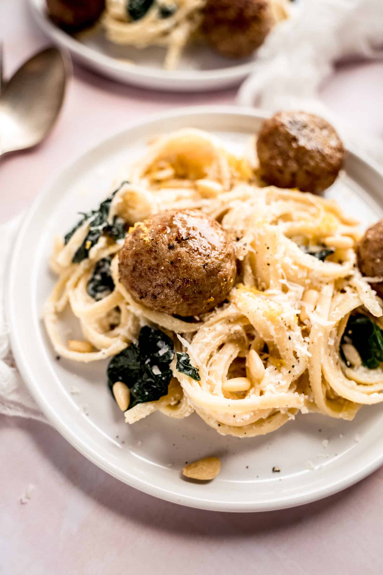 Close up shot of Lemon ricotta pasta recipe served on a white plate with creamy linguine tossed in ricotta sauce, wilted spinach, toasted pine nuts, and topped with juicy meatballs, styled in a warm, bright food photography shot.
