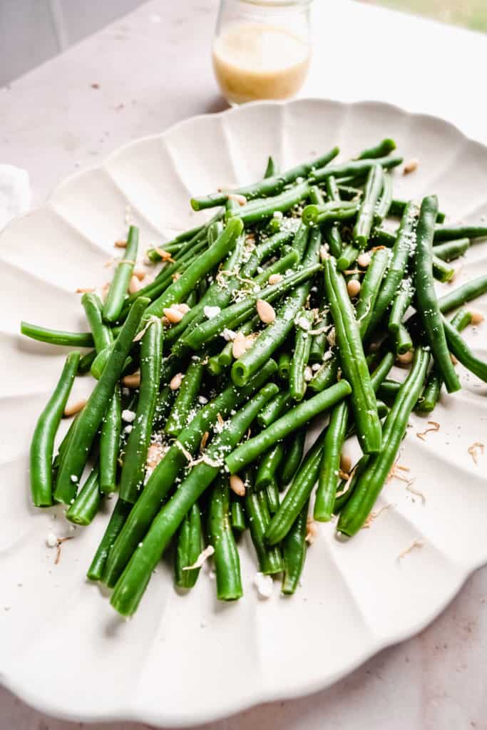 Close up shot of green bean salad garnished with pine nuts, goat cheese and dressing on scalloped platter