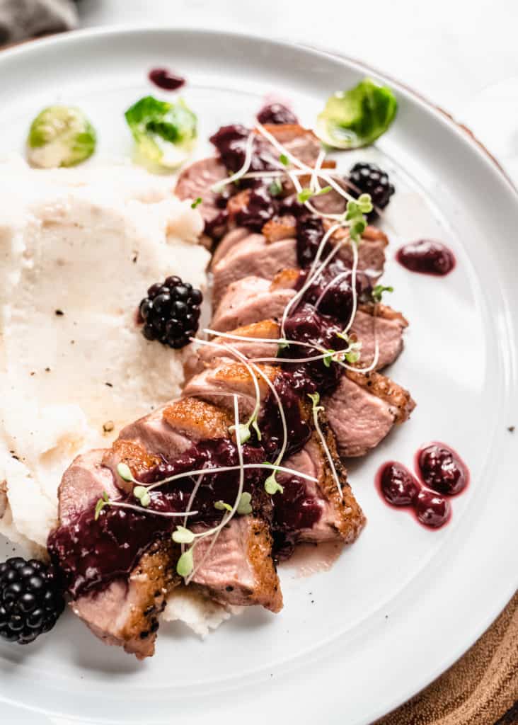 pan seared duck breast with blackberry sauce poured over top, fresh blackberries on the plate and mashed potatoes on the side. Green brussels sprout leaves are on the plate.