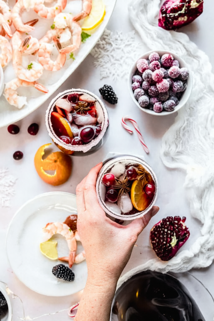A hand reaches in to pick up a sugar-rimmed glass of Christmas sangria filled with ice, cranberries, citrus slices, and star anise, surrounded by shrimp cocktail, sugared cranberries, pomegranate, and festive holiday decorations on a bright winter-themed table.