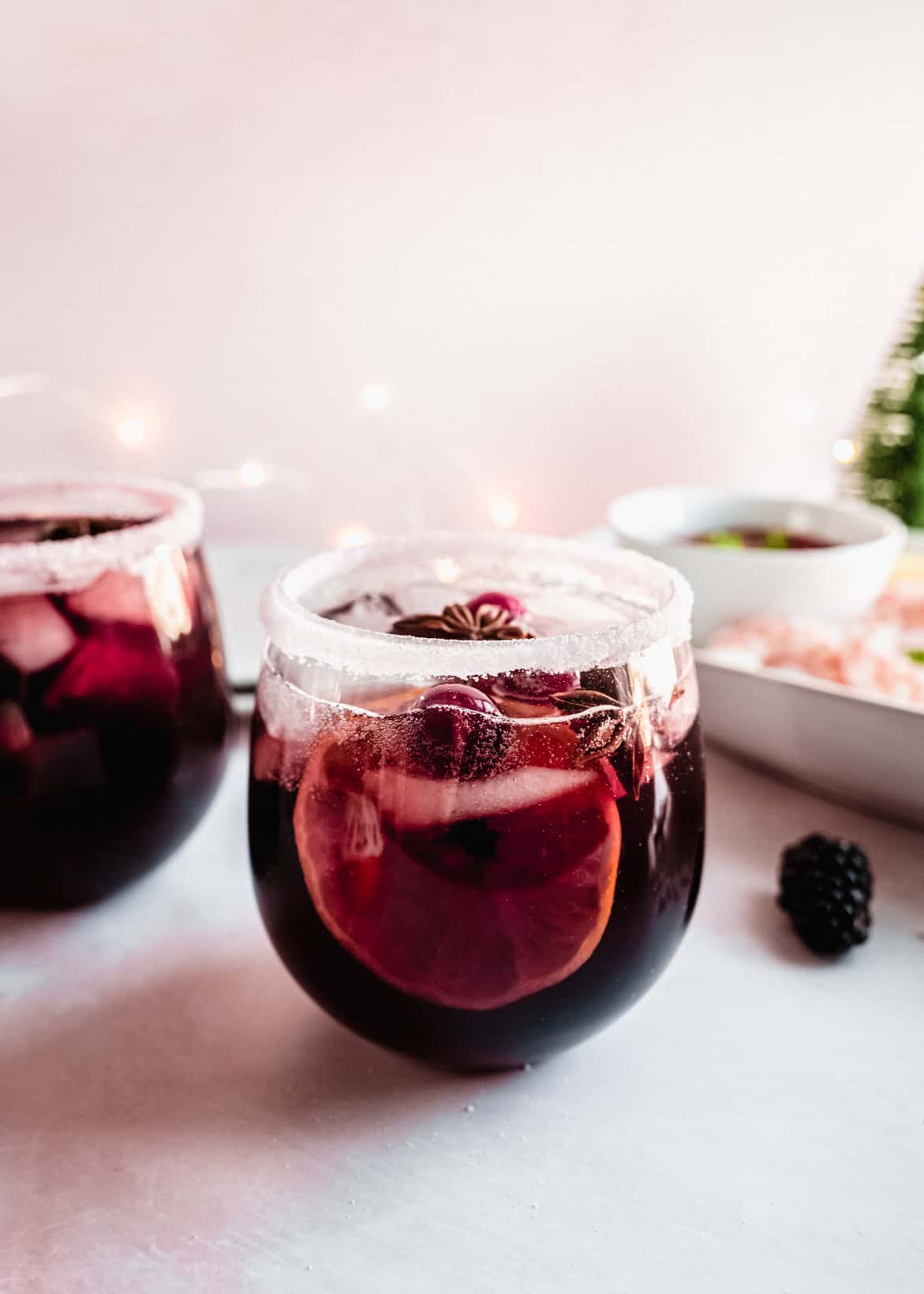 Glass of Christmas sangria filled with deep red wine, citrus slices, cranberries, and a star anise garnish, served in a sugar-rimmed stemless glass with soft holiday lights glowing in the background.