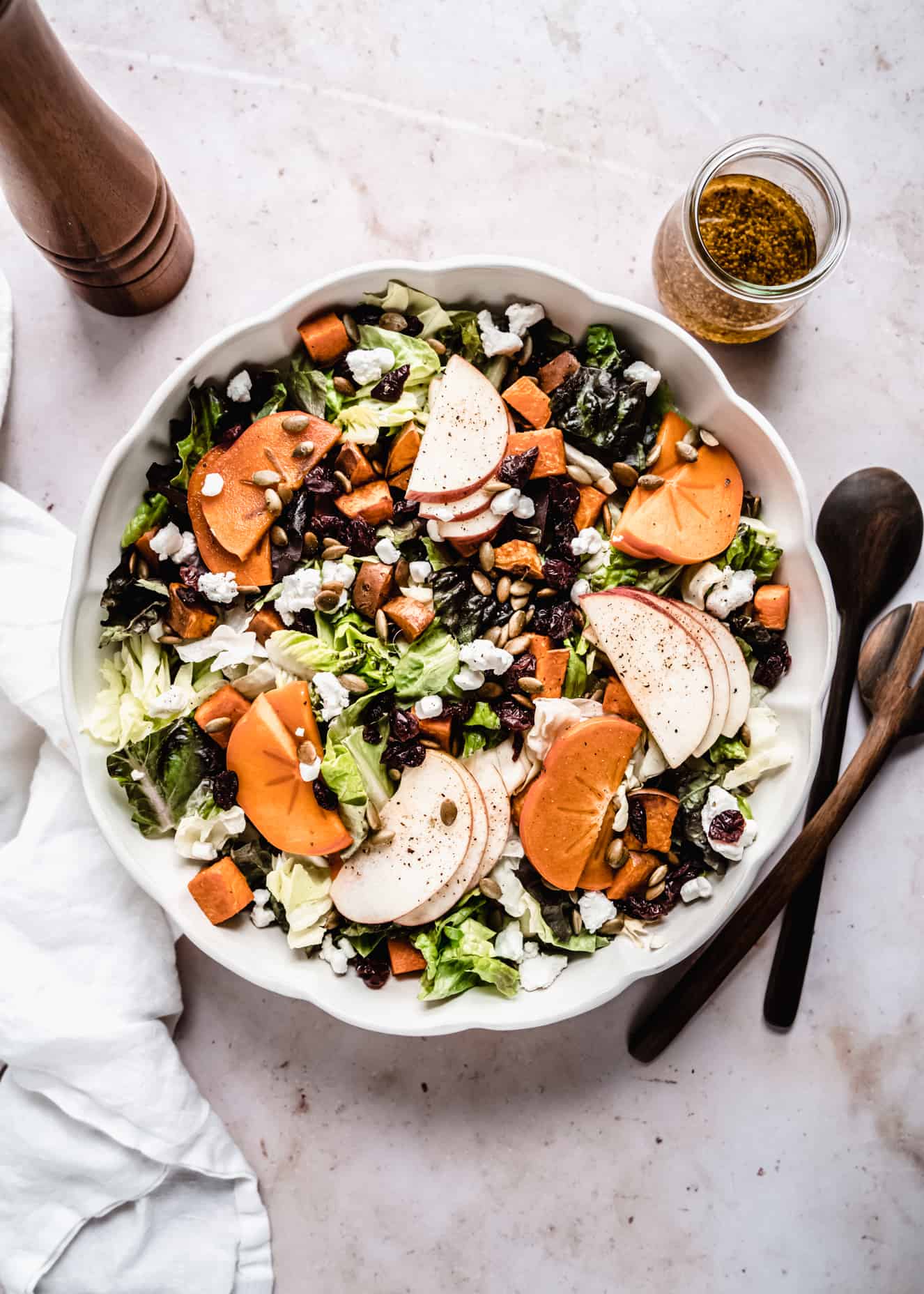 quick fresh persimmons salad in scalloped bowl with wood serving utensils next to bowl. Salad dressing, pepper grinder and white napkin are next to salad bowl.