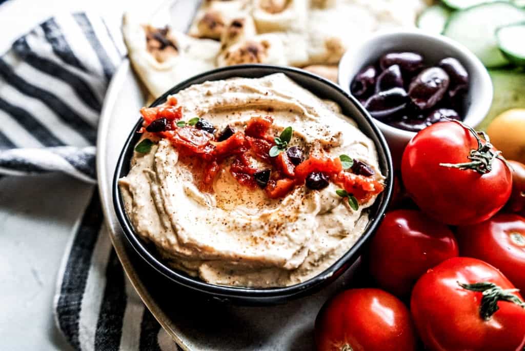 overhead shot of sundried tomato dip in a small bowl on a large platter with fresh tomatoes, cucumbers, and pita bread