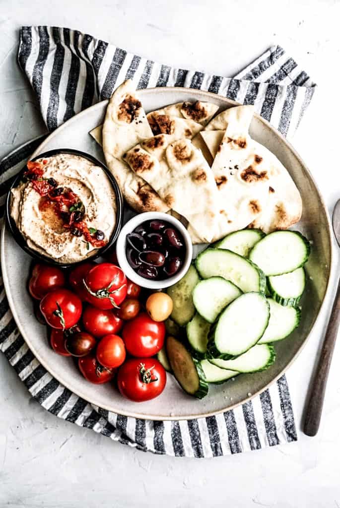 overhead shot of sundried tomato dip in a small bowl on a large platter with fresh tomatoes, cucumbers, and pita bread