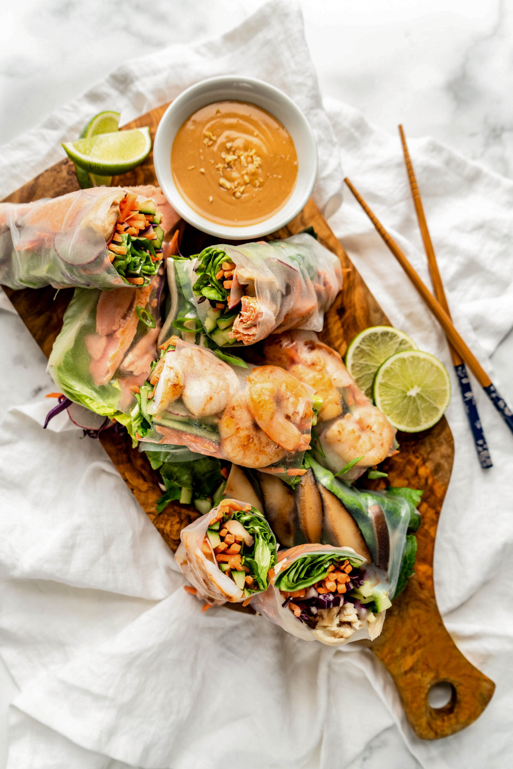 Warm, light and bright overhead photo of fresh spring rolls arranged on a wooden board with a bowl of creamy peanut sauce for spring rolls, garnished with chopped peanuts and served alongside lime wedges and chopsticks on a white marble background.