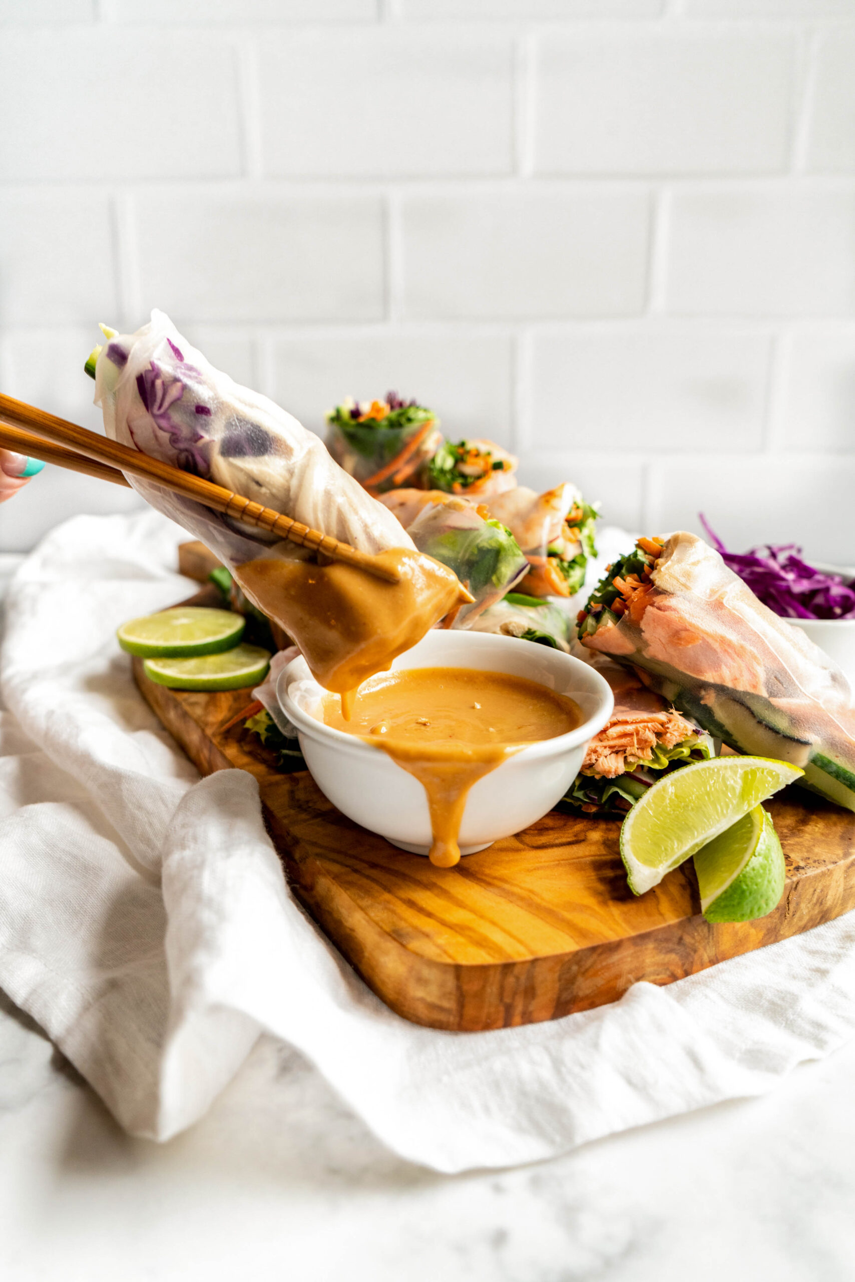 Warm, light and bright photo of fresh spring rolls being dipped into creamy peanut sauce for spring rolls, with chopsticks holding a roll above a small bowl as the sauce drips down, served on a wooden board with lime wedges and colorful veggies in the background.
