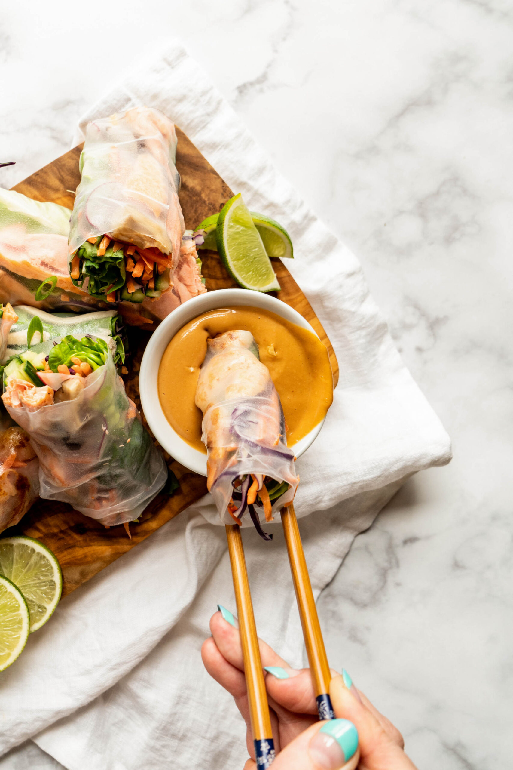 Warm, light and bright photo of fresh spring rolls being dipped into creamy peanut sauce for spring rolls, with chopsticks holding a roll above a small bowl as the sauce drips down, served on a wooden board with lime wedges and colorful veggies in the background.
