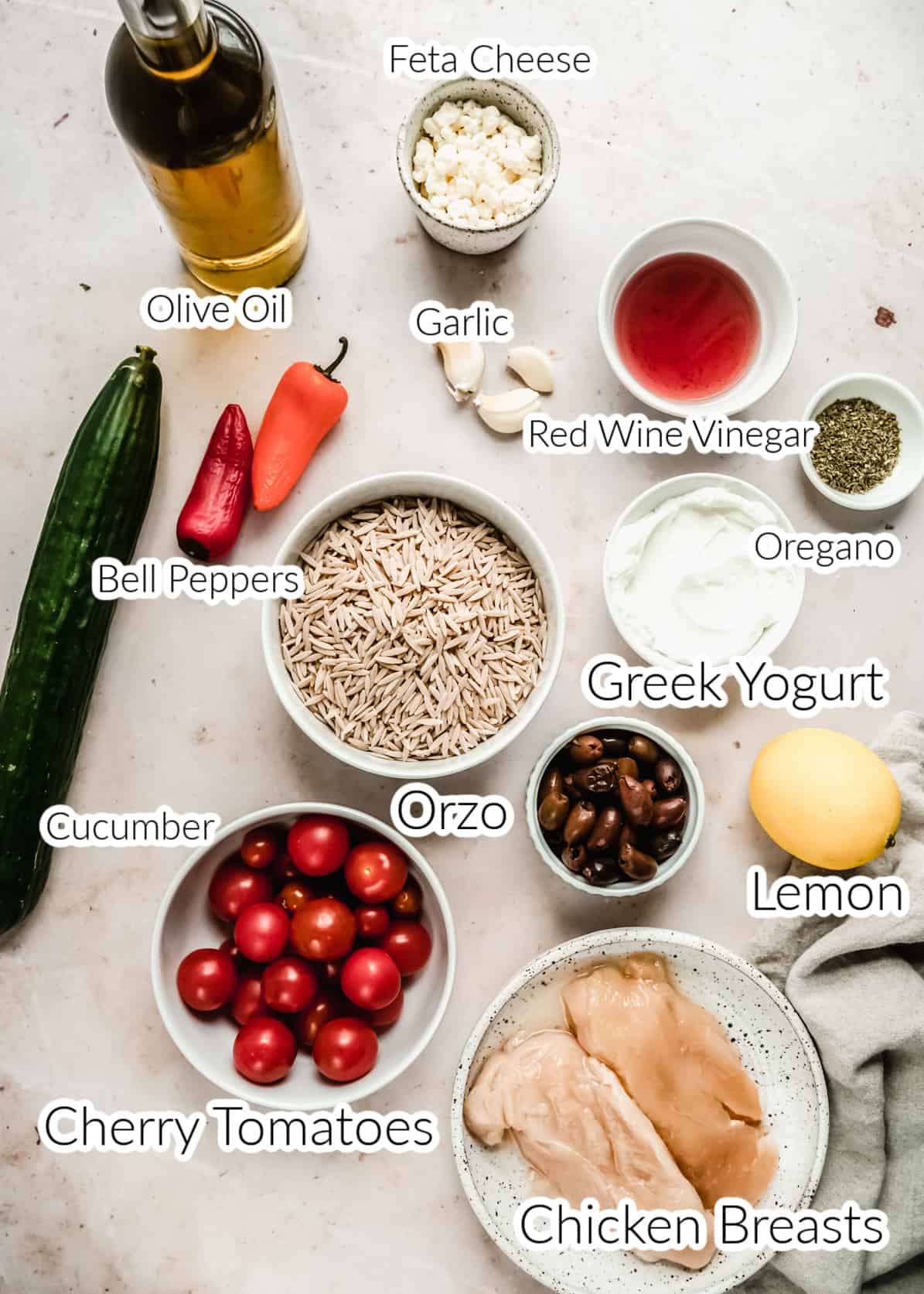 Overhead view of fresh ingredients arranged on a light marble surface for Greek chicken bowls, including raw chicken breasts, orzo, cherry tomatoes, cucumber, bell peppers, kalamata olives, feta cheese, Greek yogurt, lemon, garlic, oregano, olive oil, and red wine vinegar in individual bowls. Bright, colorful Mediterranean components styled for meal prep.