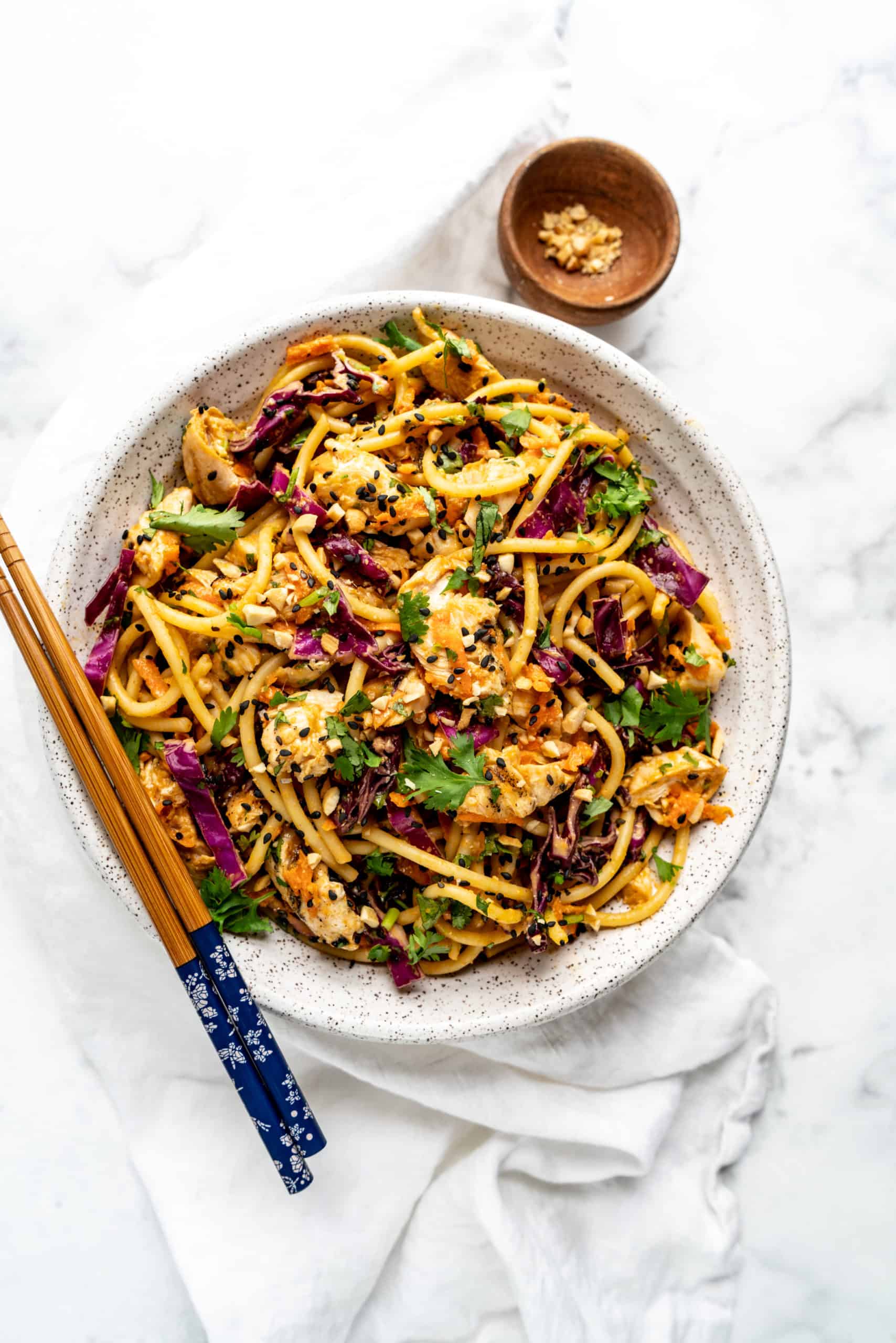Overhead view of a bowl of Peanut Thai noodle salad tossed with spaghetti noodles, shredded red cabbage, carrots, chopped cilantro, tender chicken, and black sesame seeds, served with chopsticks on a white marble surface.