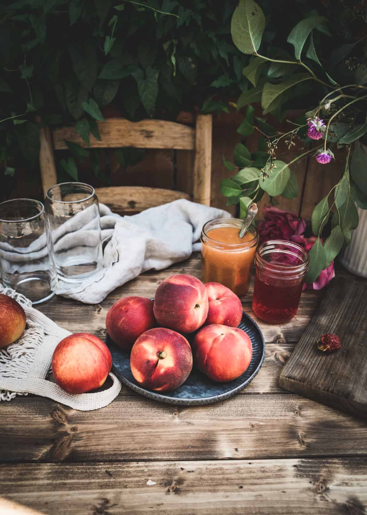 Whole fresh peaches on metal plate on wood table. Peach puree and peach simple syrup are in glass jars behind the plate, and empty glasses are on the table. Greenery surrounds the table.