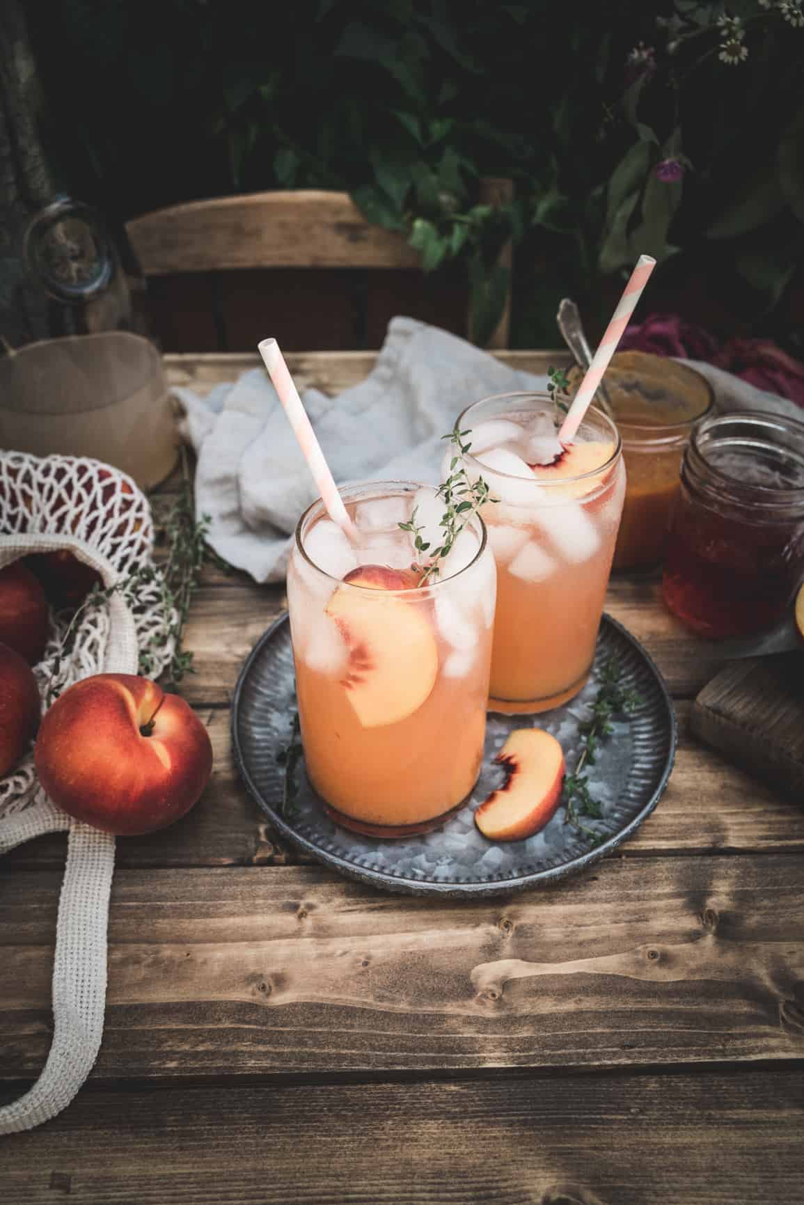 Whole fresh peaches on metal plate on wood table. Peach puree and peach simple syrup are in glass jars behind the plate, and empty glasses are on the table. Greenery surrounds the table.