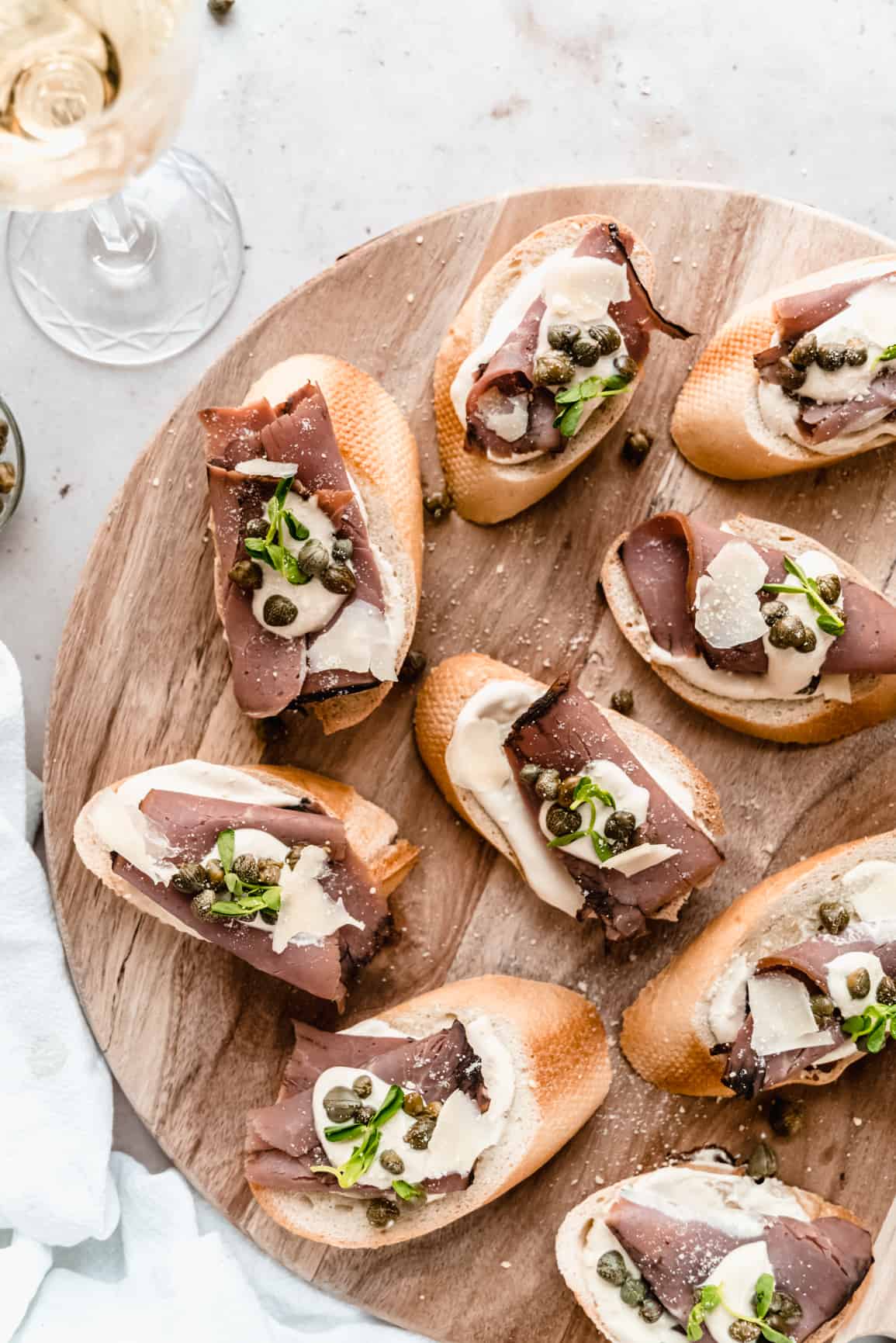 overhead shot of roast beef crostini garnished with horseradish dijon sauce, capers and microgreens on a wood serving platter, placed next to a glass of wine