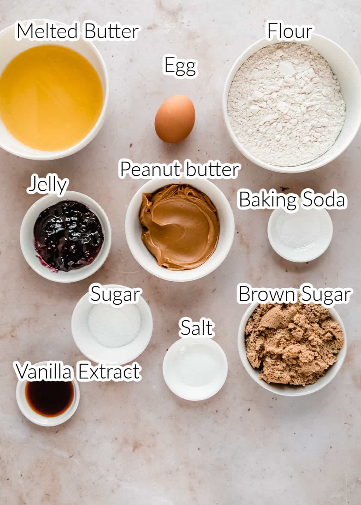 Labeled ingredients for making peanut butter and jelly cookies arranged on a marble surface, including bowls of peanut butter, jelly, flour, brown sugar, granulated sugar, melted butter, baking soda, salt, vanilla extract, and a whole egg.