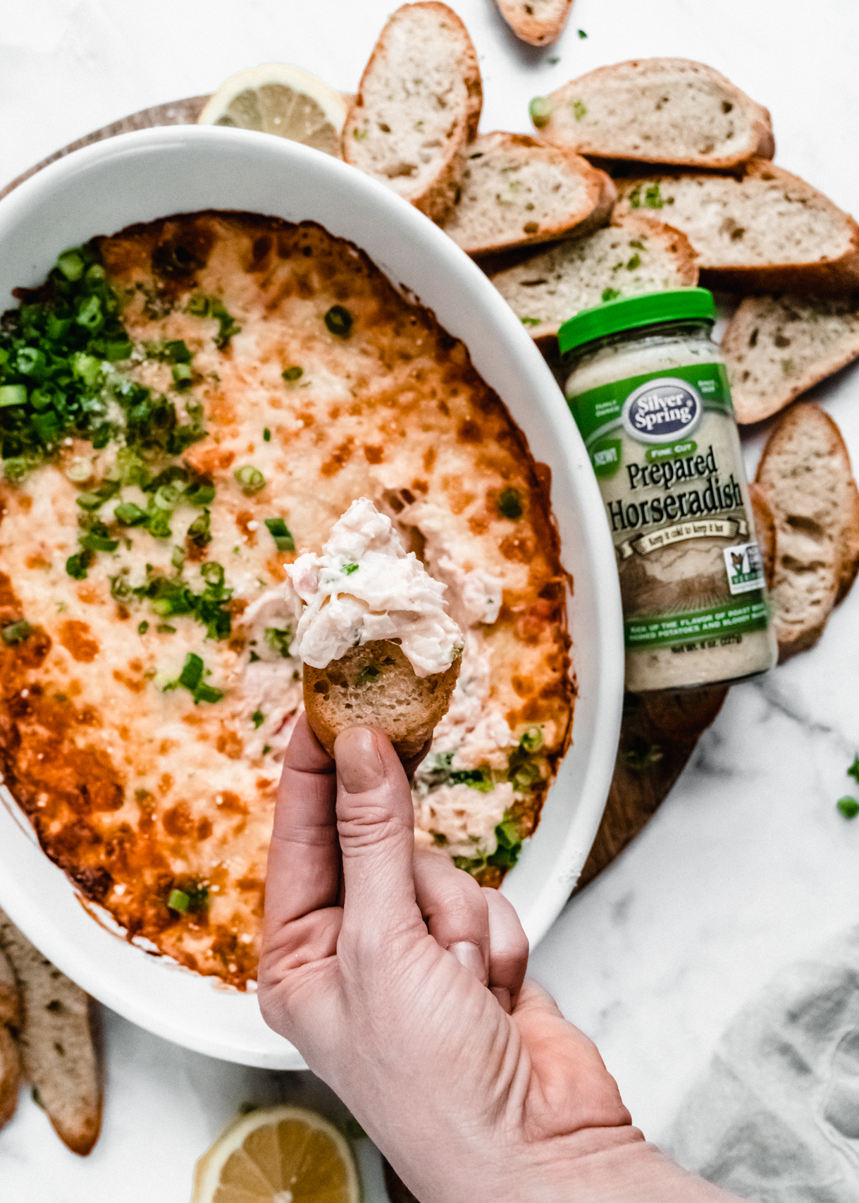a hand dipping a toasted baguette slice scooping out baked crab rangoon dip in baking dish 