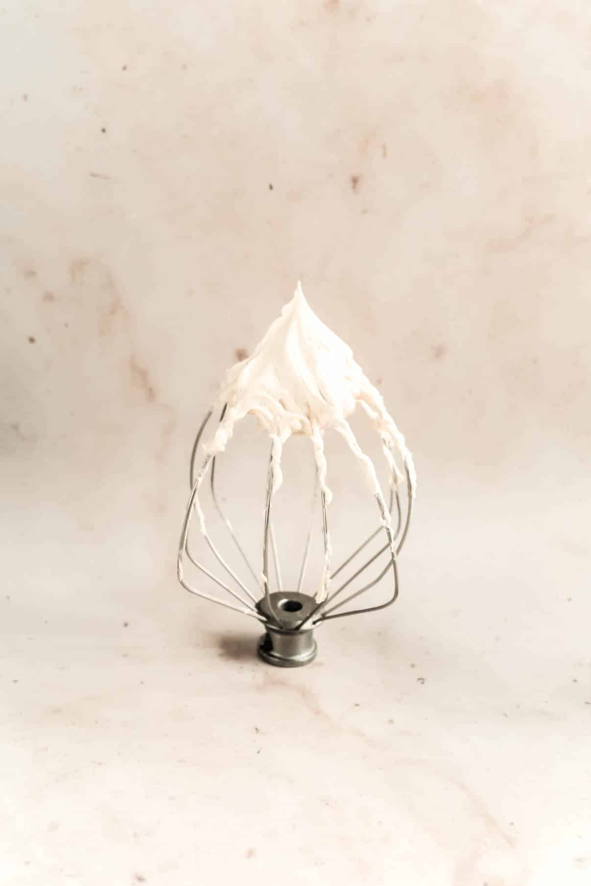 A mixer whisk attachment coated with a peak of creamy frosting, set against a light marble background, showing the frosting preparation for a gingerbread snack cake.”