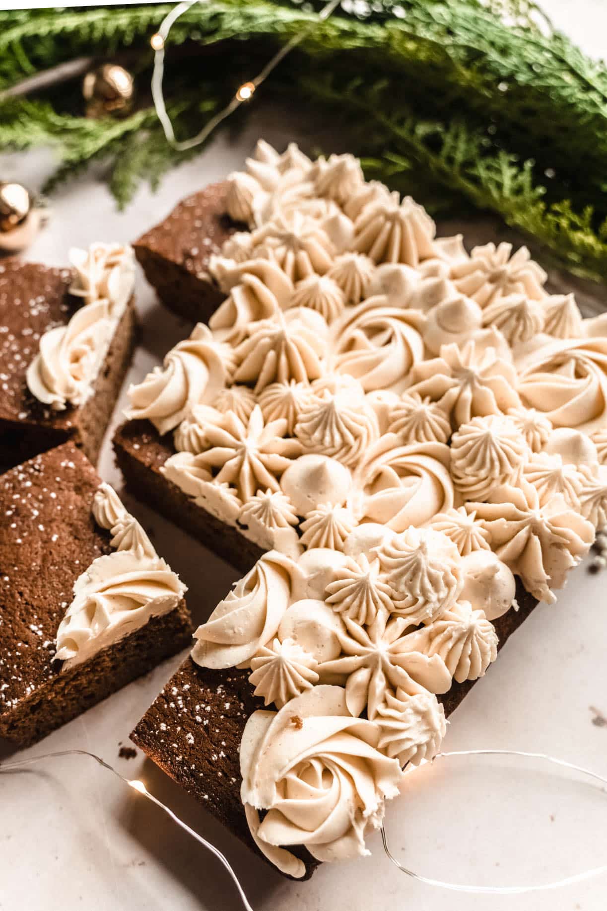 Close-up of a gingerbread snack cake cut into squares and decorated with piped swirls and stars of creamy frosting, surrounded by holiday greenery and twinkle lights.