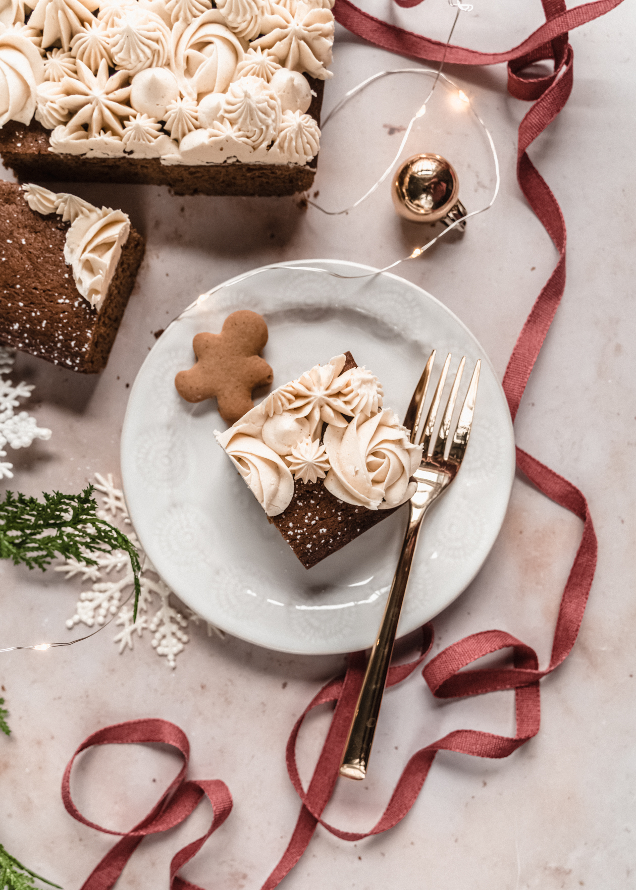 square piece of gingerbread snack cake frosted on small white plate with gold fork and tiny gingerbread man. Plate is surrounded by red ribbon and greenery