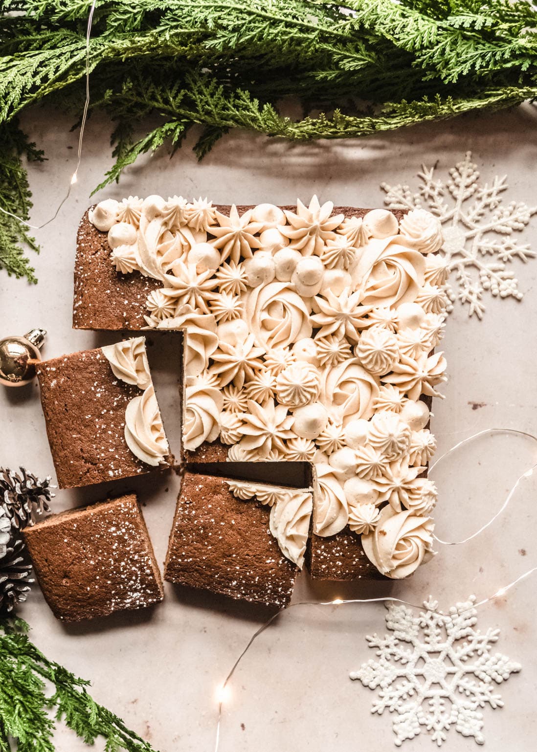 A gingerbread snack cake decorated with intricate swirls and stars of buttercream frosting is cut into squares, displayed on a festive surface with greenery, pinecones, snowflakes, and twinkle lights