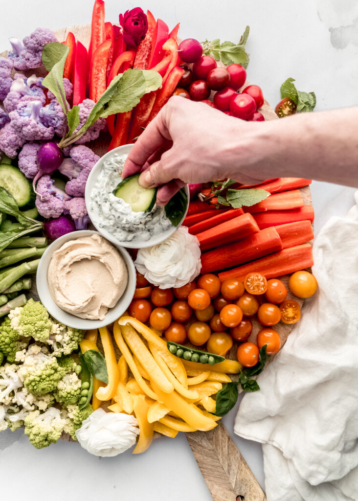 a hand dipping a cucumber slice into the dip bowl in the middle of the crudité platter