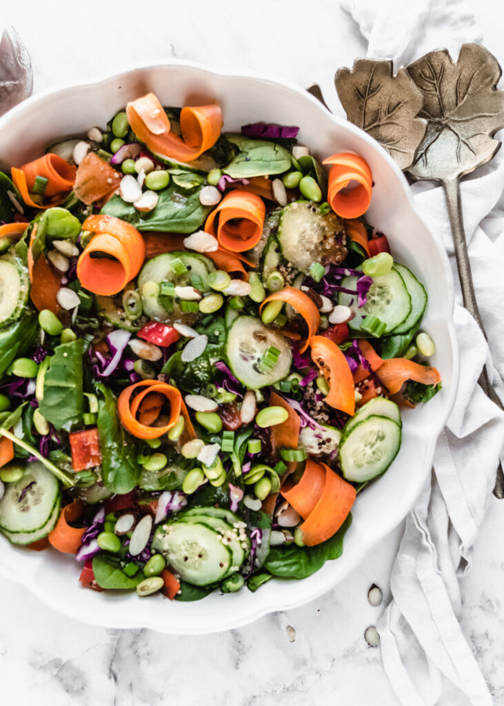overhead shot of asian edamame salad in scalloped bowl with aged gold salad tongs to the right of the bowl on top of white napkin